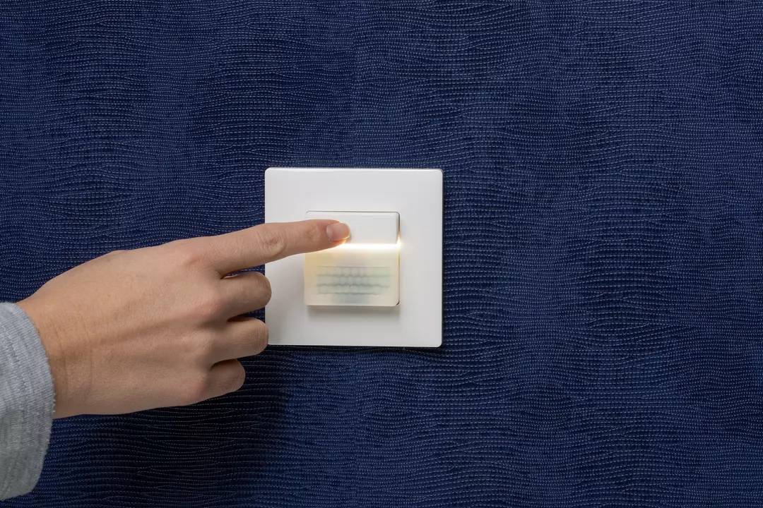 Eine Hand drückt einen Lichtschalter an einer blauen Wand. Der Schalter leuchtet leicht auf und zeigt eine Berührung in Aktion.