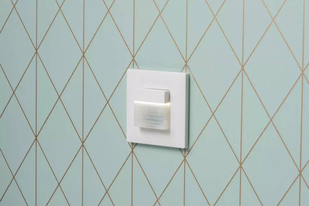 Schalter mit Bewegungssensor an einer blau-gemusterten Wand. Der Sensor leuchtet leicht, bereit zur Erfassung von Bewegungen.