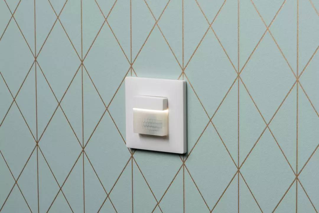 Ein weißer Lichtschalter an einer Wand mit blauem Rautenmuster. Der Schalter hat ein kleines LED-Licht in der Mitte.