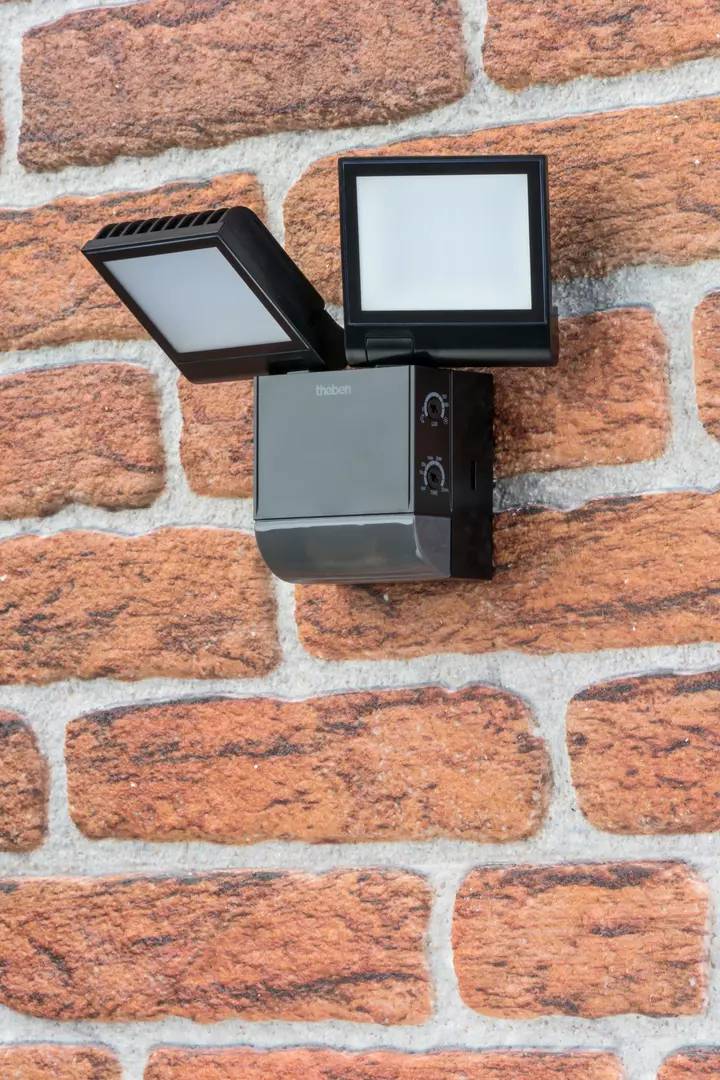 Eine schwarze Außenlampe mit Bewegungssensor ist an einer Mauer aus roten Ziegelsteinen montiert.