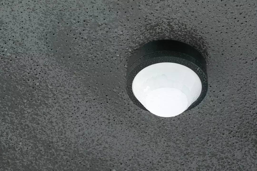 Eine schwarze Deckenlampe mit rundem Design hängt an einer grauen Betonoberfläche. Sie beleuchtet den Raum minimalistisch.