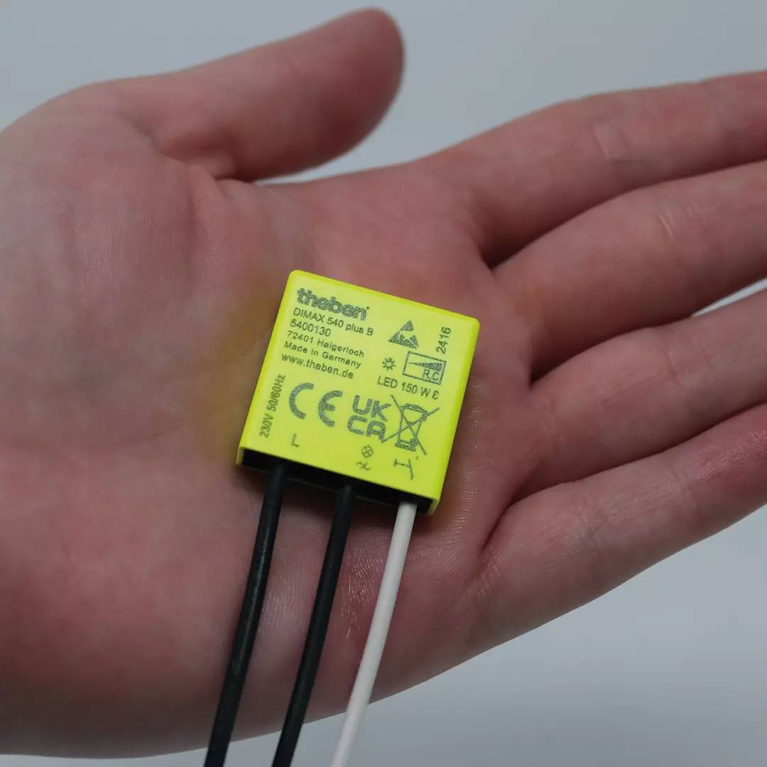 Eine Hand hält einen kleinen, gelben elektronischen Baustein mit der Aufschrift 'theben' und technischen Details. Gerät hat drei Kabel.