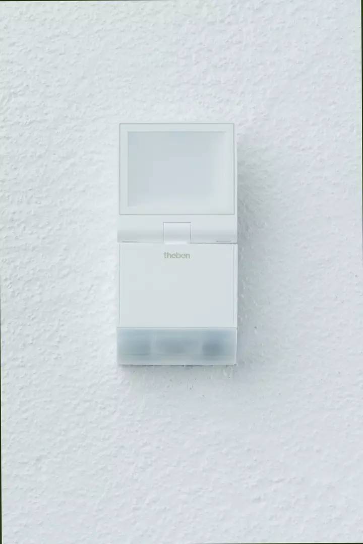Ein weißer Bewegungsmelder mit dem Logo 'theben' an einer weißen Wand montiert, wahrscheinlich für die Überwachung eines Bereichs.