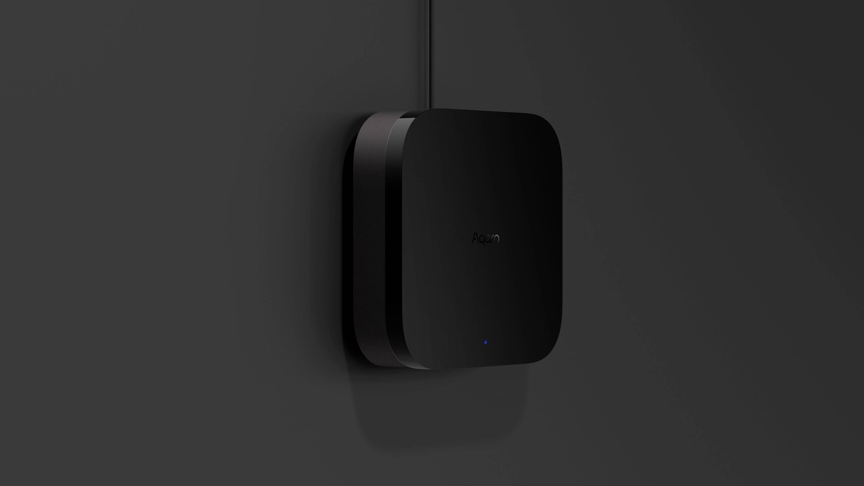 Ein schwarzer, rechteckiger Router mit abgerundeten Ecken hängt an einer dunkelgrauen Wand. Ein blaues Licht leuchtet unten.