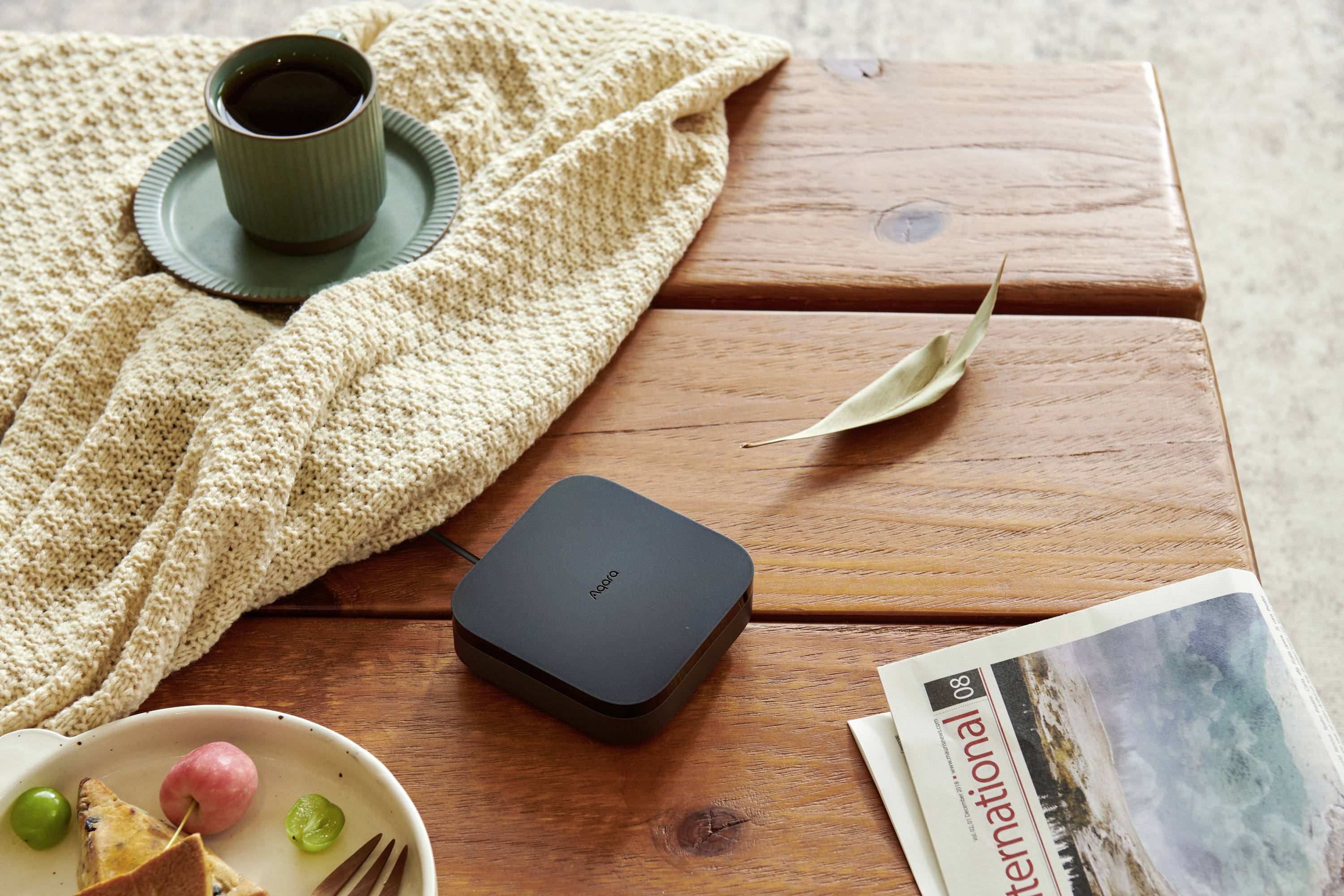 Holztisch mit Funkladegerät, Kaffeetasse, Zeitung und Obstteller darauf. Entspannende Atmosphäre mit neutralen Farben.