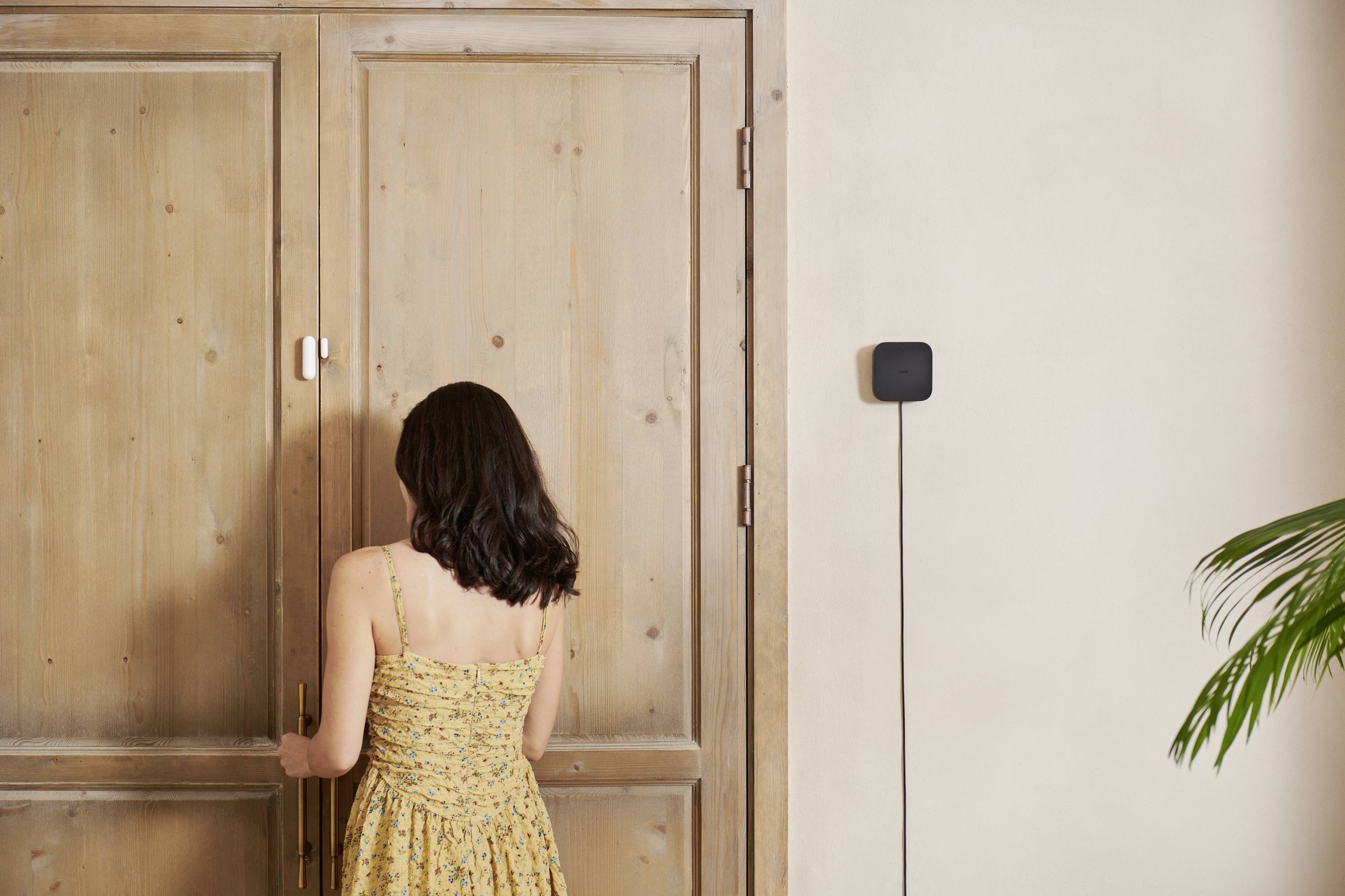 Eine Frau in einem gelben Kleid steht vor einer geschlossenen Holztür. An der Wand hängt ein kleines schwarzes Gerät mit einem Kabel.