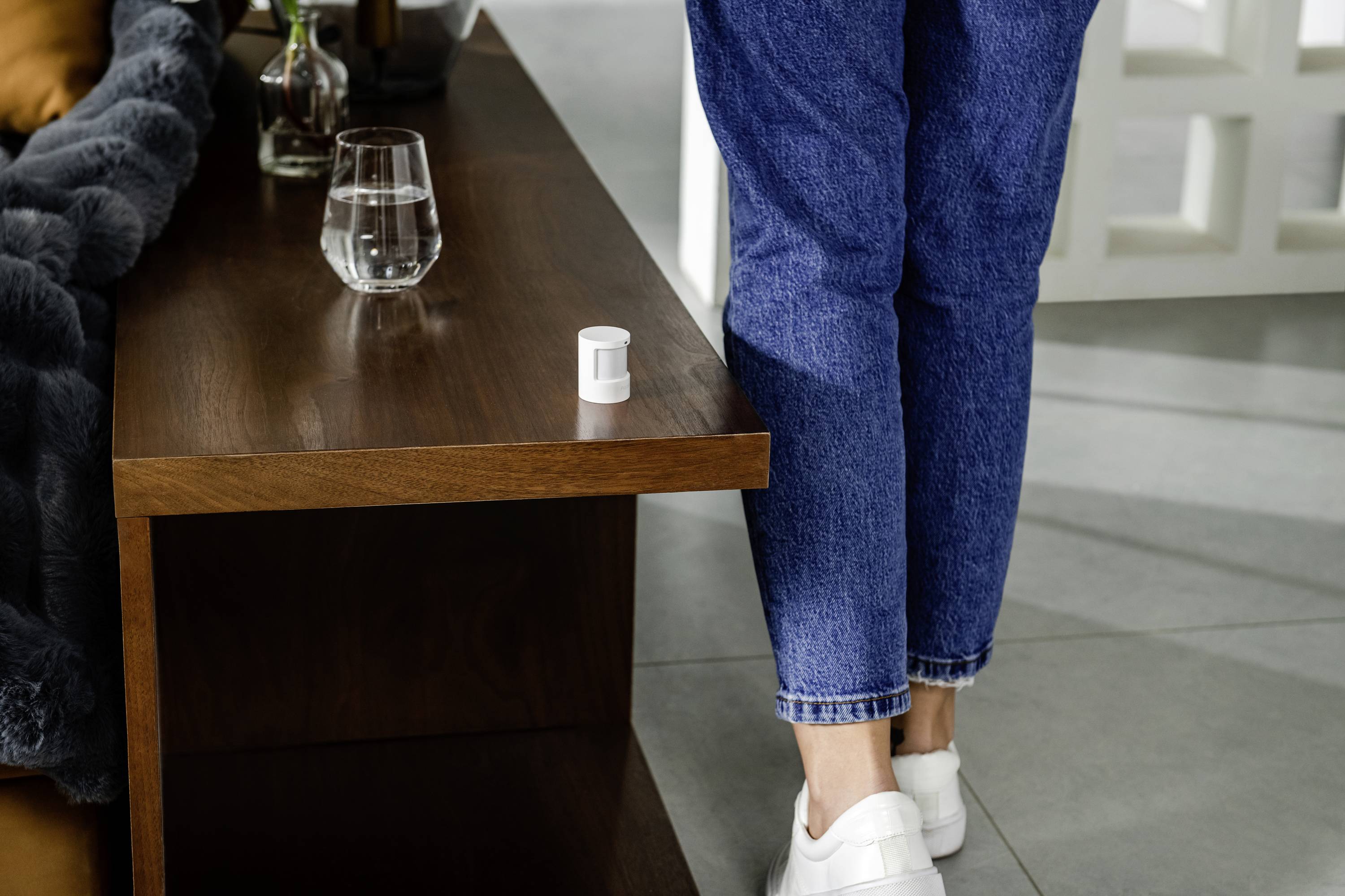 Eine Person in blauen Jeans und weißen Schuhen steht neben einem Holztisch. Auf dem Tisch steht ein Glas Wasser und ein kleiner Behälter.