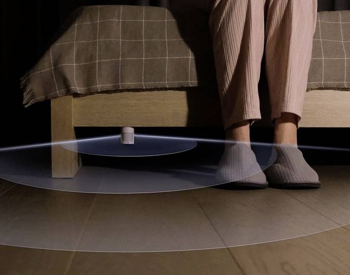 Eine Person sitzt auf einer Bettkante, darunter ein kleines Gerät. Ein digitaler Radius ist auf den Boden projiziert, zeigt Geräteabdeckung an.