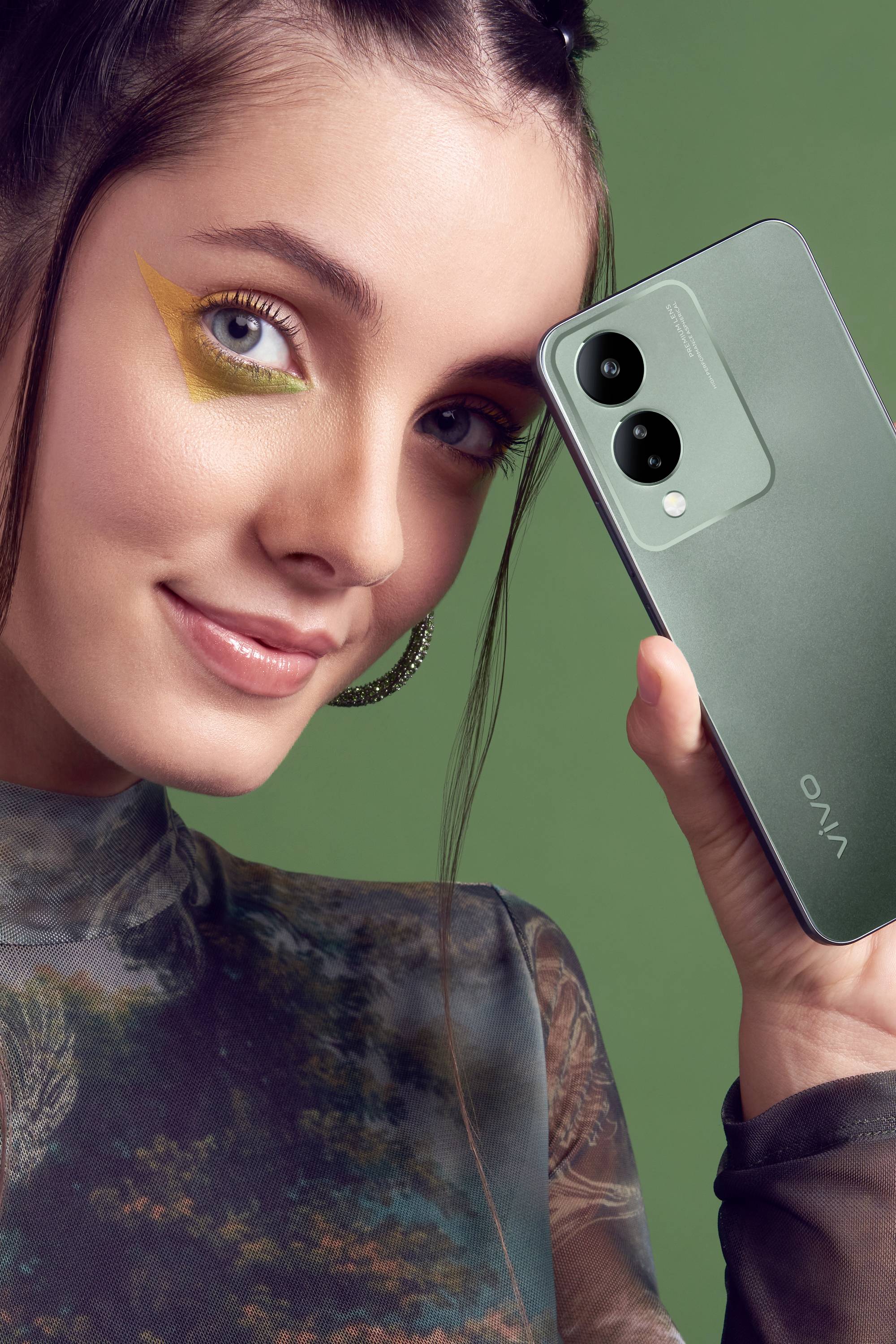 Eine Frau hält ein Smartphone von Vivo vor einem grünen Hintergrund. Sie lächelt und trägt auffälliges gelbes Augen-Make-up.