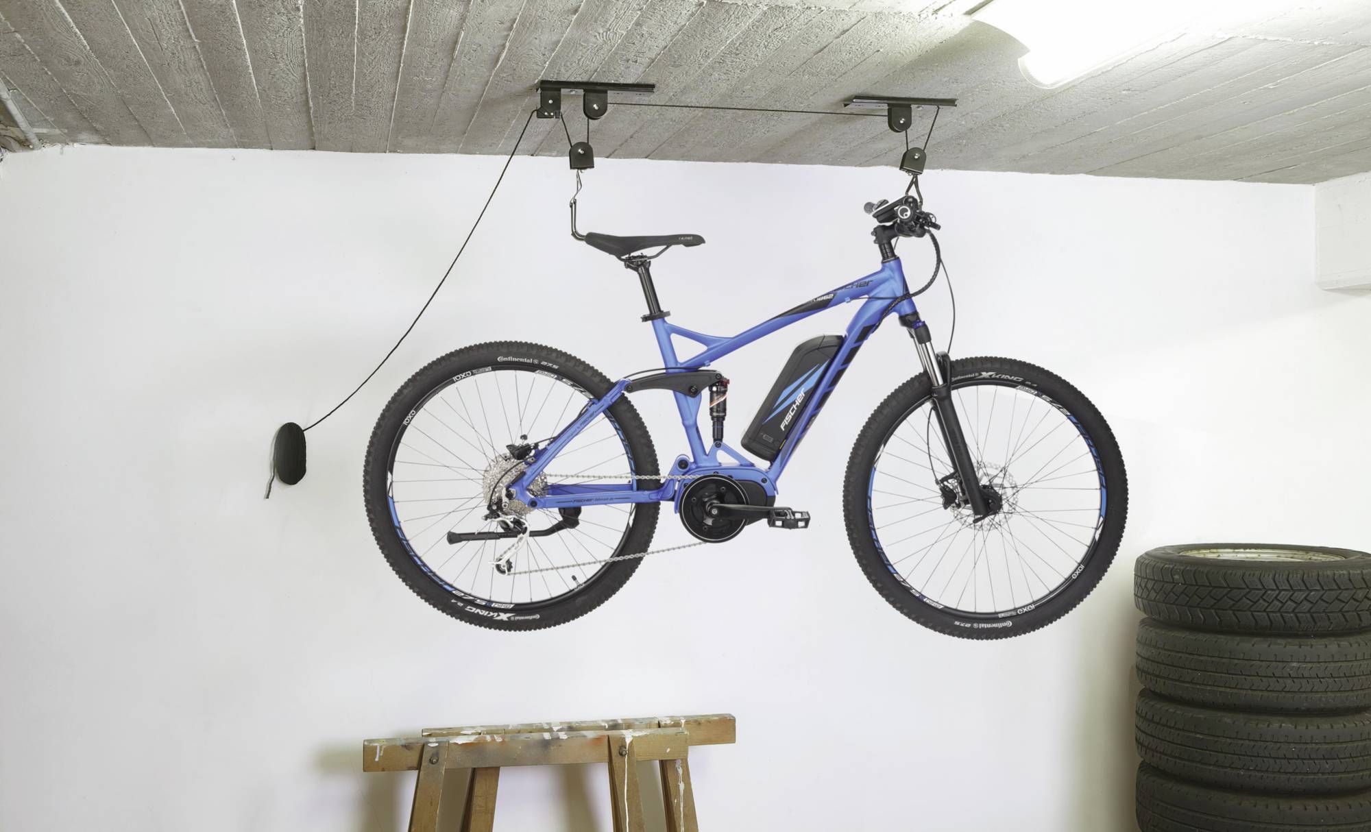 Ein blaues Fahrrad hängt an einer Deckenhalterung in einer Garage. Rechts sind gestapelte Reifen zu sehen, darunter eine hölzerne Werkbank.
