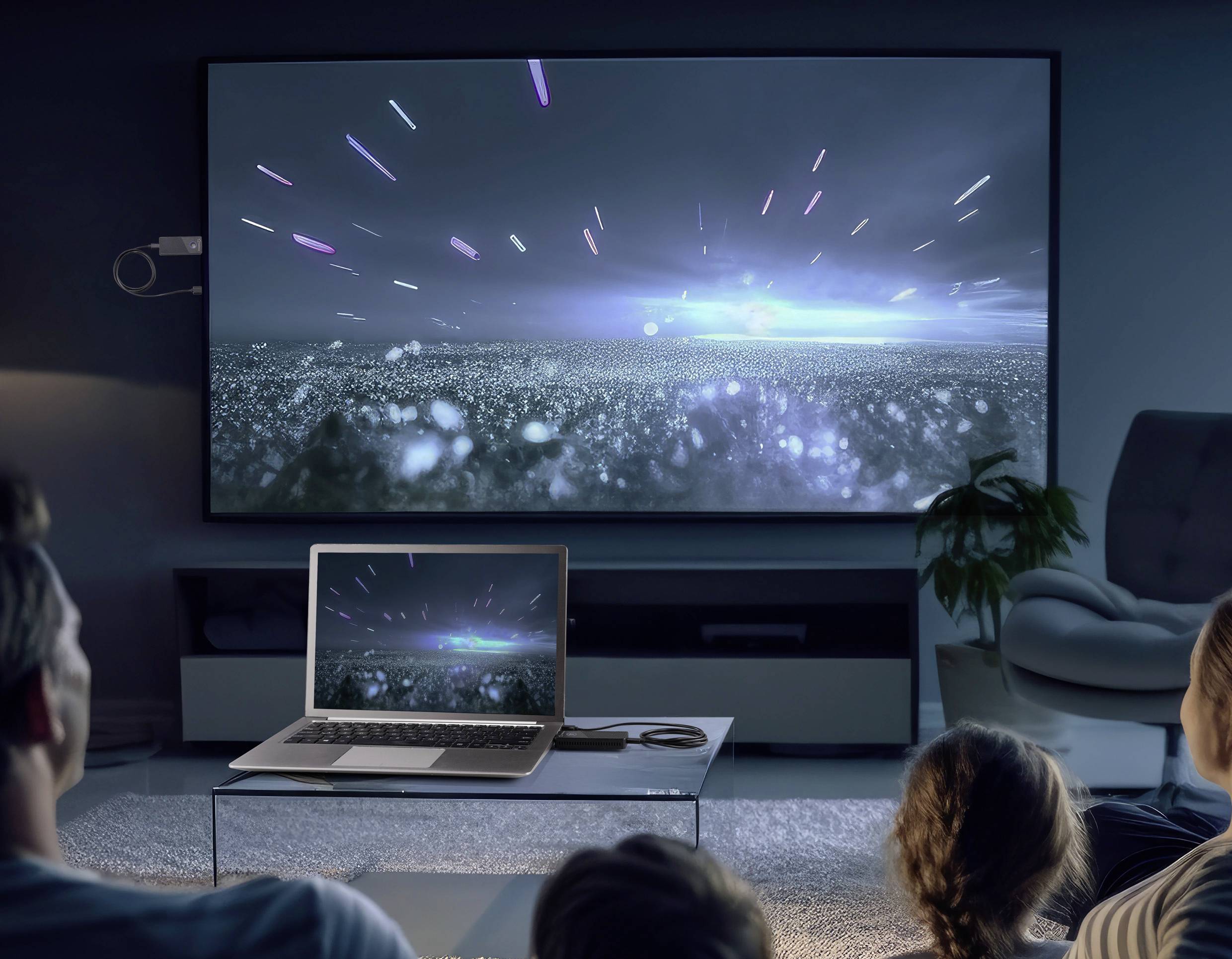 Eine Familie sitzt in einem abgedunkelten Raum und schaut eine Science-Fiction-Szene mit Lichtstrahlen auf einem großen Fernseher, während ein Laptop auf dem Tisch dasselbe Bild zeigt.