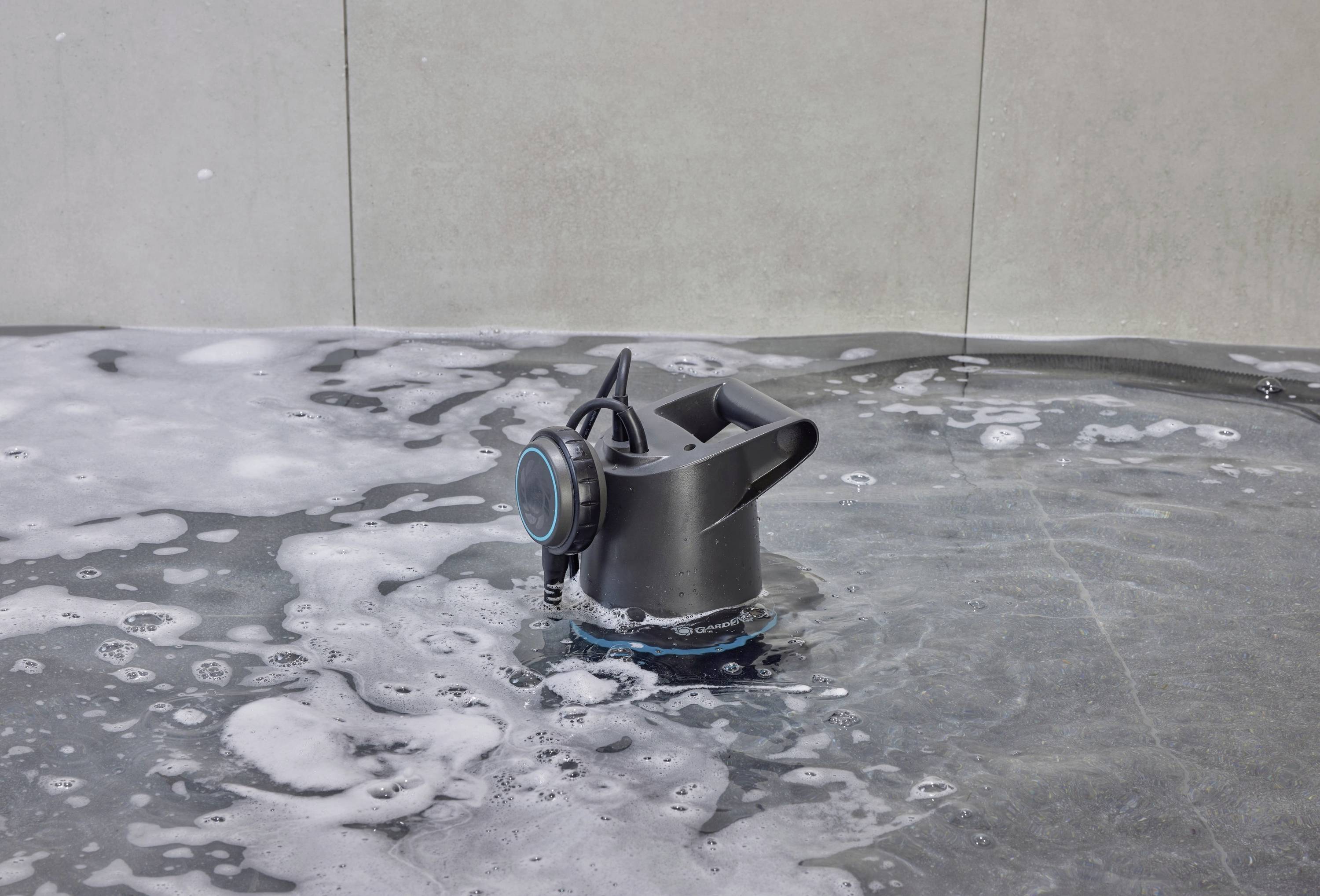 Ein schwarzer Wasserlautsprecher schwimmt in einem Becken mit Schaum. Der Lautsprecher hat ein rundes, leuchtendes Display.