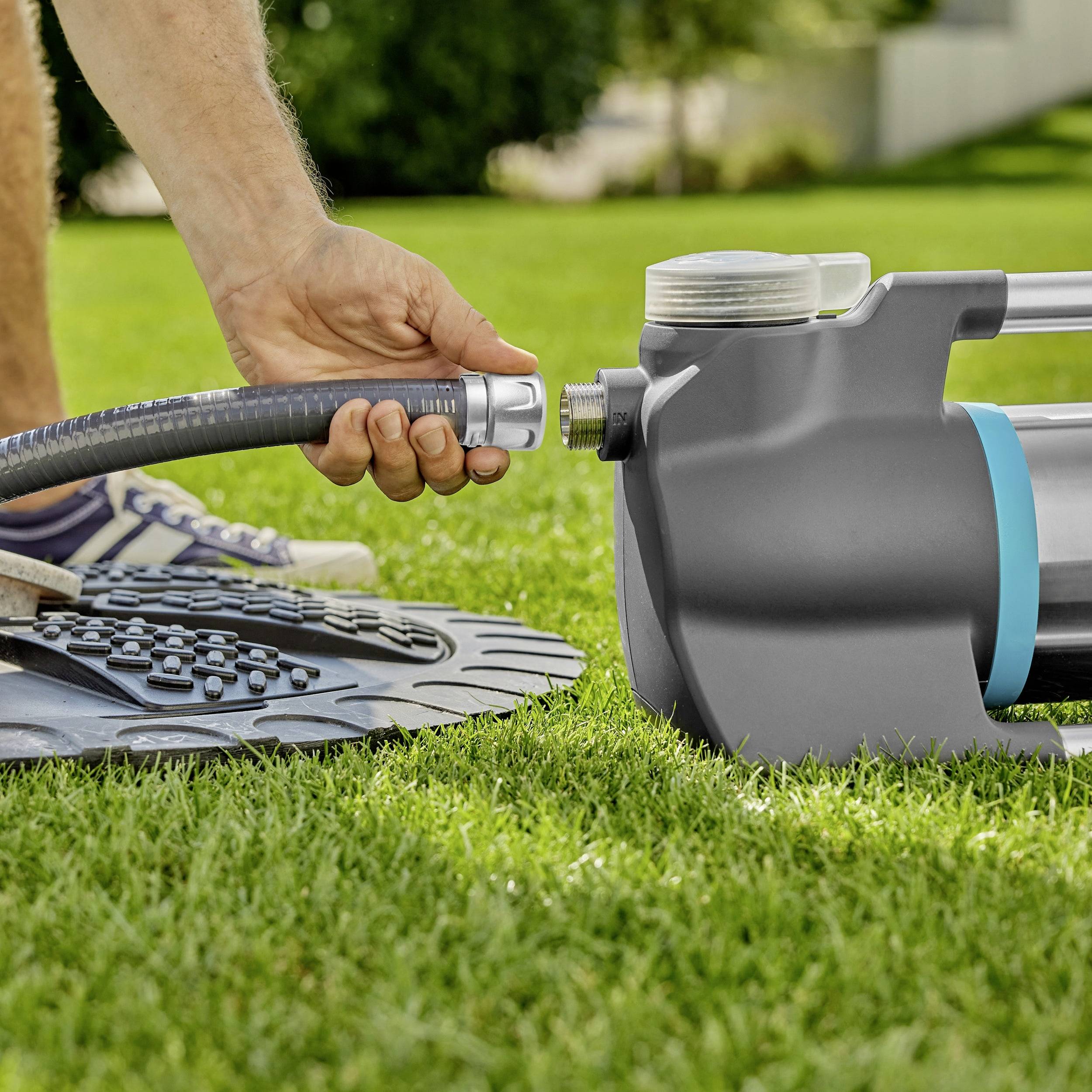 Person verbindet einen Gartenschlauch mit einer Wasserpumpe auf einem Rasen. Die Szene zeigt die Vorbereitung zur Gartenbewässerung.