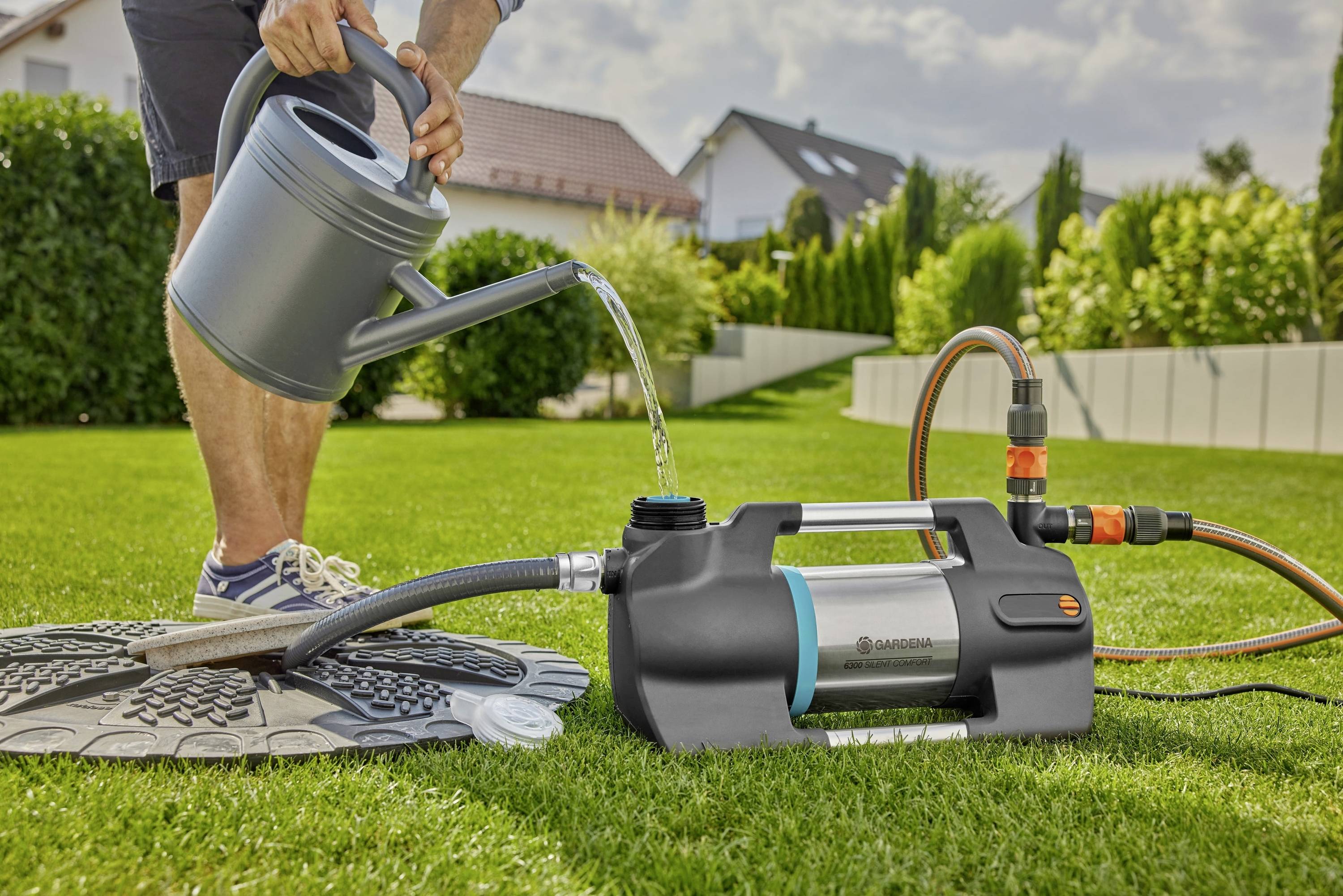 Eine Person gießt Wasser aus einer Gießkanne in eine Gartenpumpe auf einem Rasen. Im Hintergrund sind Häuser und Pflanzen zu sehen.