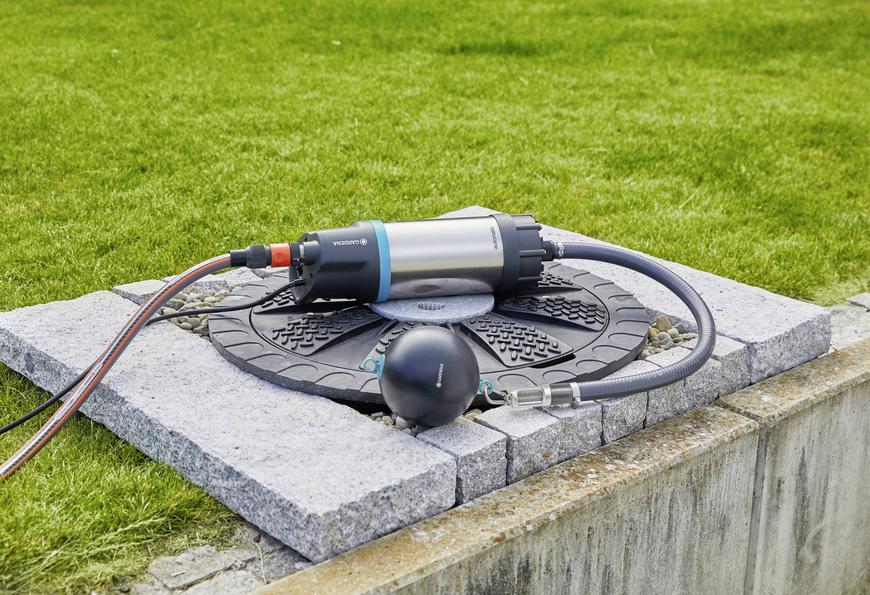Eine silberne Wasserpumpe mit Schläuchen liegt auf einem Brunnendeckel im Gartenbereich, umgeben von grünem Rasen.