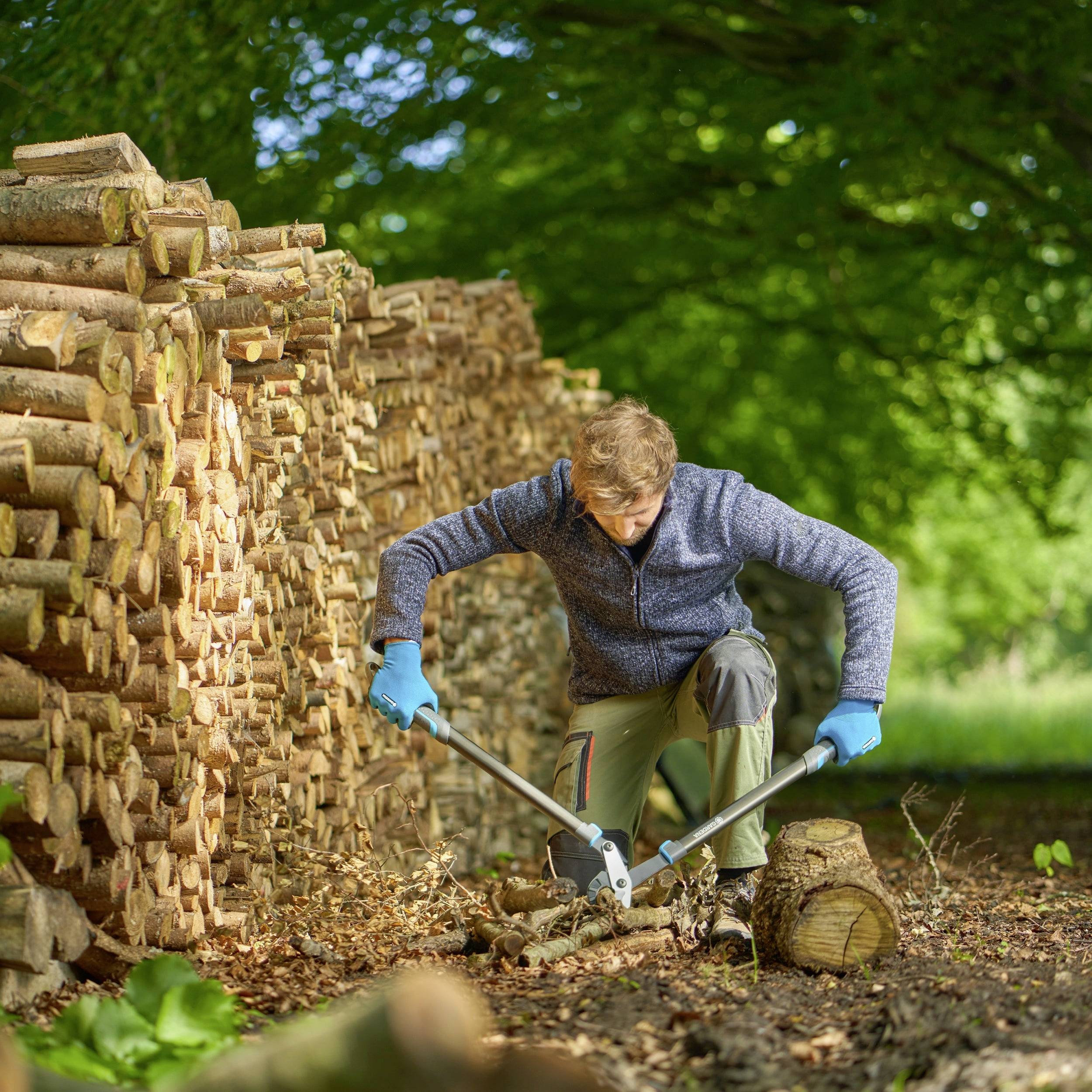Eine Person spaltet Holz mit einer Axt vor einem großen Stapel Holzscheite. Im Hintergrund ist eine grüne Wiese zu sehen.