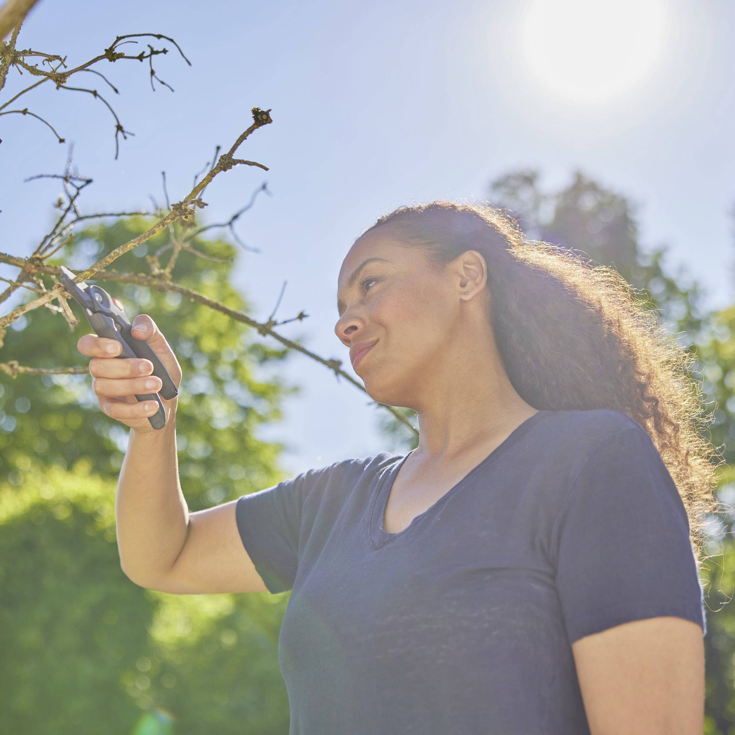 Eine Frau schneidet einen Ast mit einer Gartenschere. Im Hintergrund sind Bäume und ein klarer Himmel zu sehen.