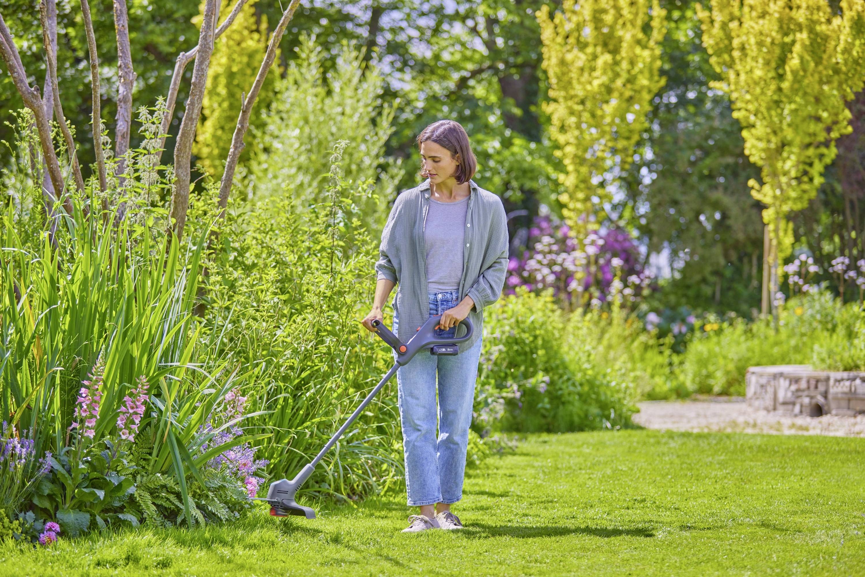 Eine Person nutzt einen elektrischen Rasentrimmer, um Rasen im Garten zu schneiden. Umgeben von blühenden Pflanzen und Bäumen.