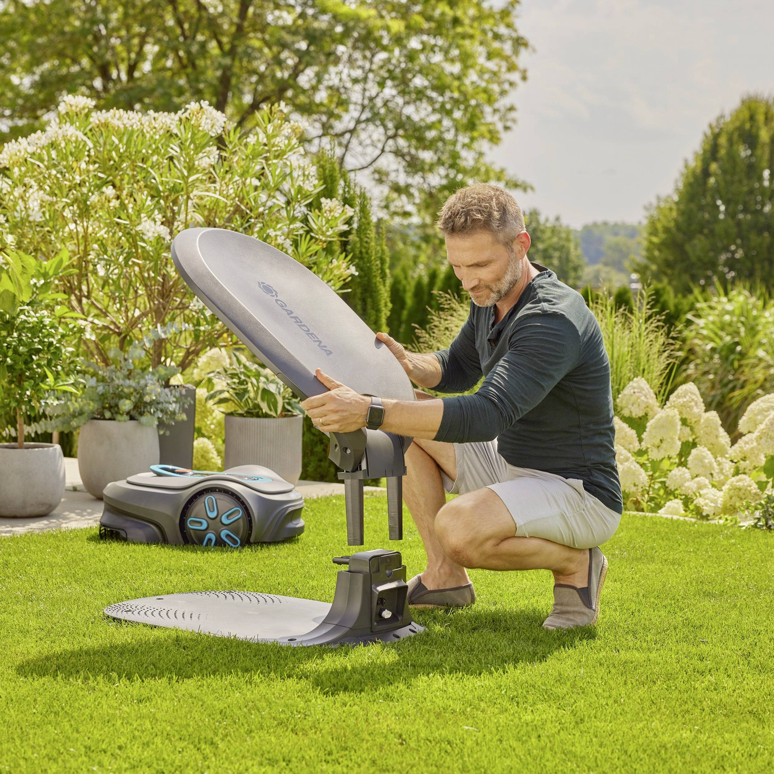 Mann montiert ein automatisches Rasenroboter-Ladestationszubehör im Garten, umgeben von grüner Vegetation und Topfpflanzen.