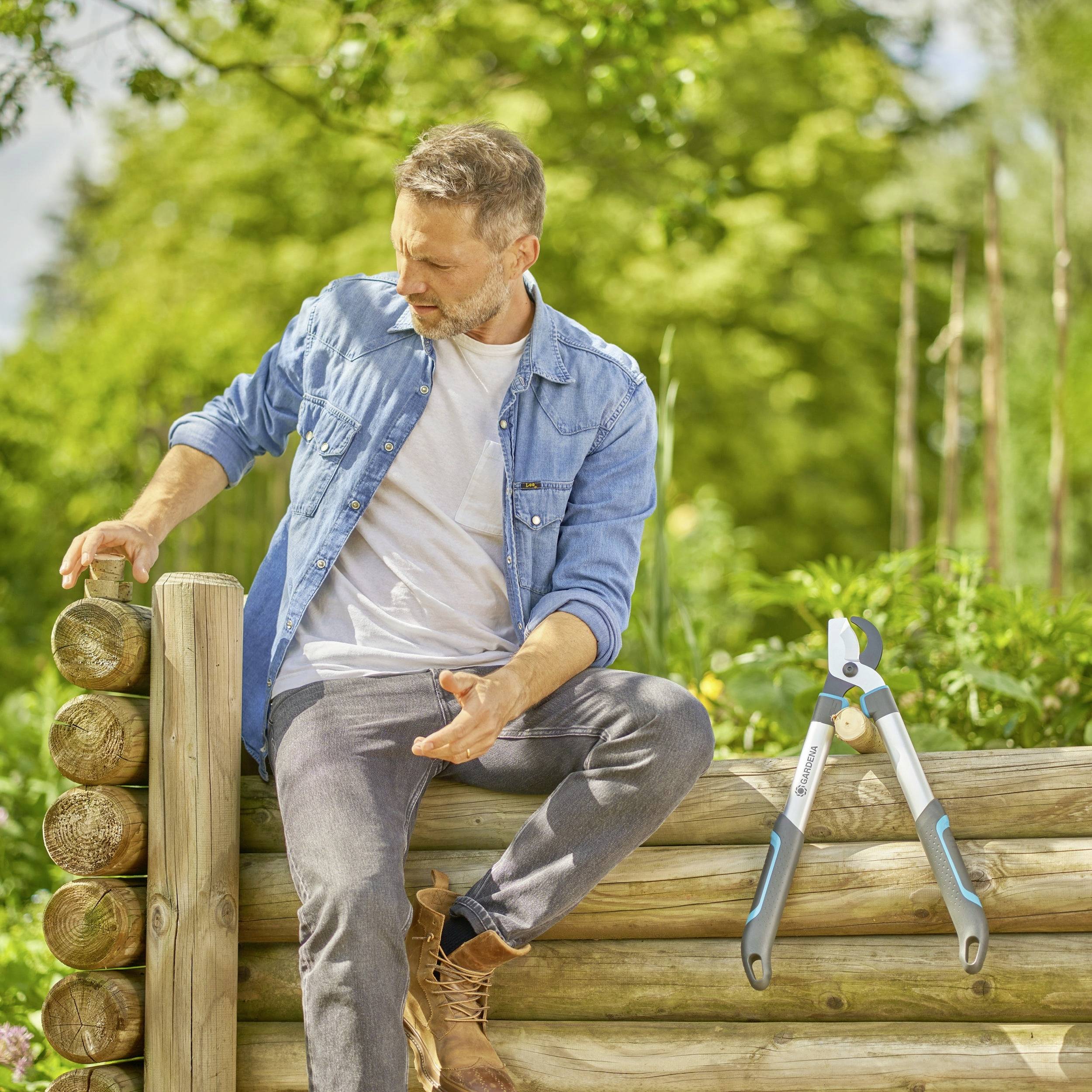 Ein Mann lehnt entspannt an einem Holzzaun in einem sonnigen Garten. Neben ihm hängt eine Gartenschere am Zaun.