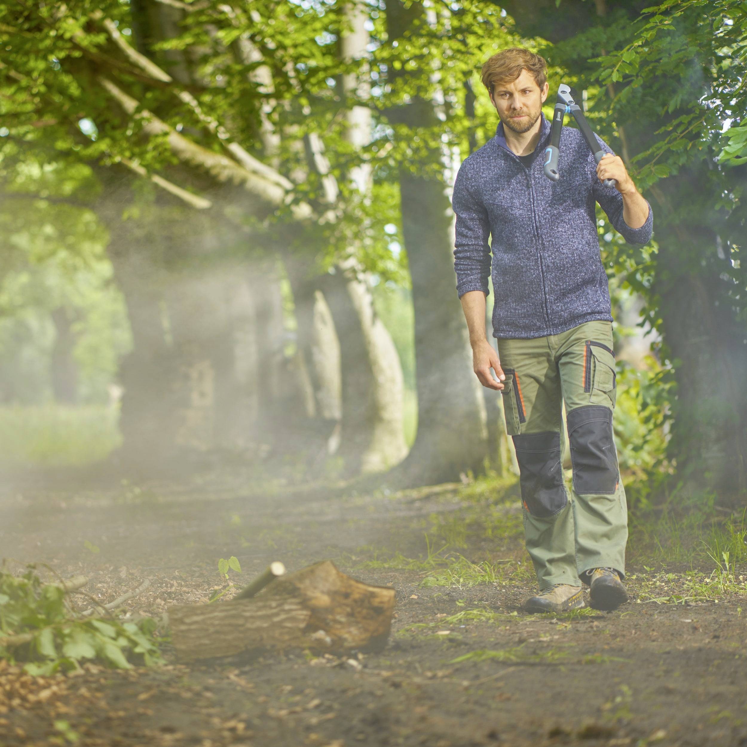 Ein Mann mit Axt spaziert durch einen Waldweg, umgeben von hohen Bäumen und Sonnenlicht, das durch das Laub fällt.