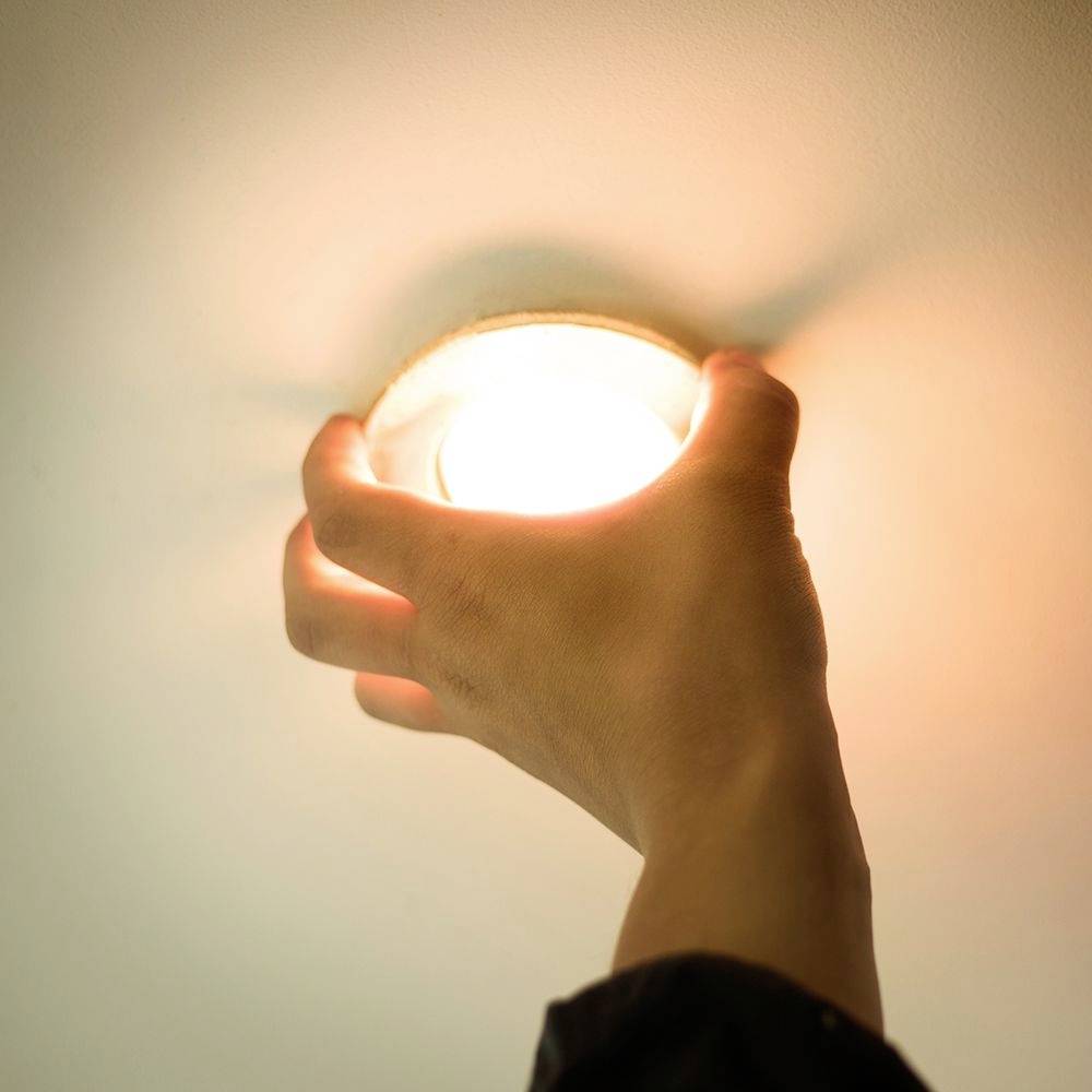 Eine Hand dreht eine Glühbirne in eine Deckenfassung, wodurch warmes Licht in den Raum abgestrahlt wird.