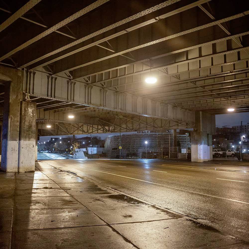 Eine nächtliche, leere Straße unter einer Brücke, beleuchtet von Straßenlaternen. Feuchte Fahrbahn reflektiert das Licht.