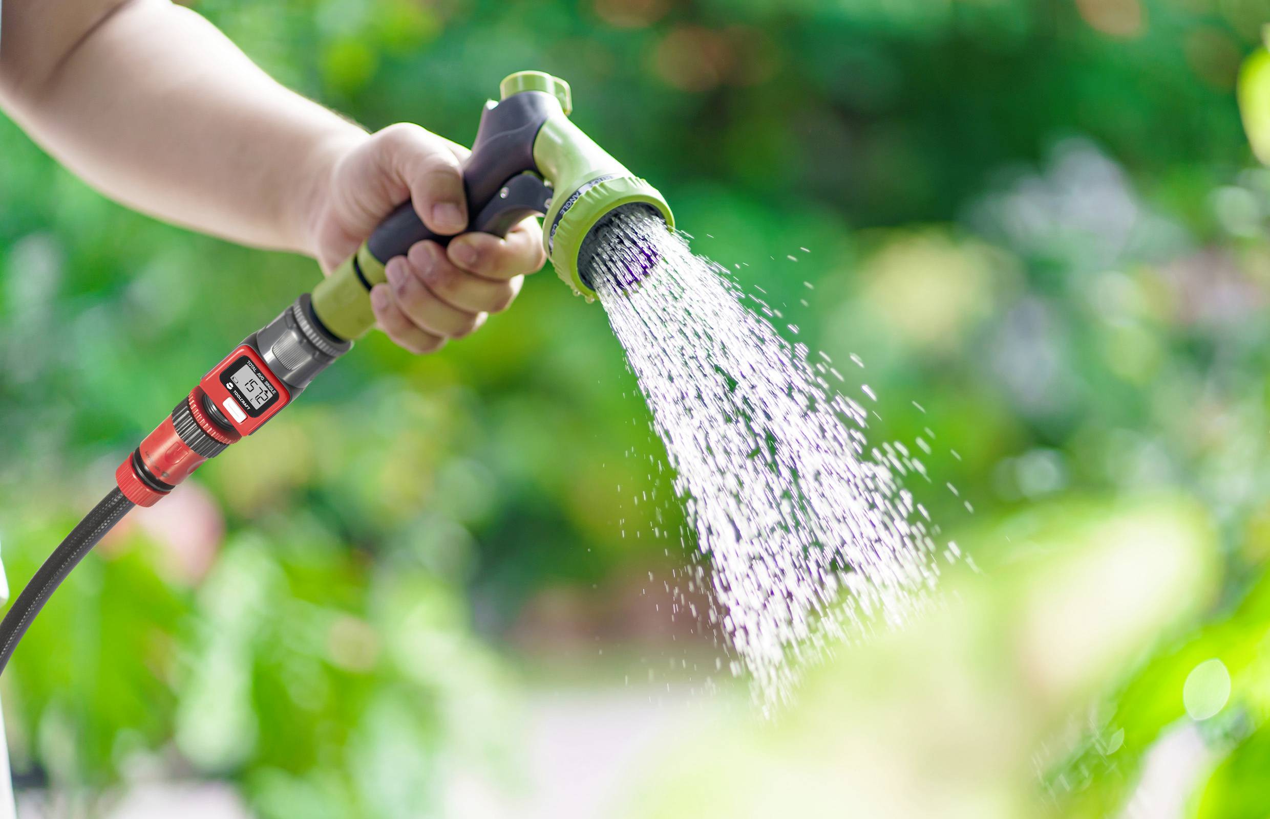 Eine Hand hält einen Gartenschlauch, aus dem Wasser spritzt, mit unscharfem Grün im Hintergrund.