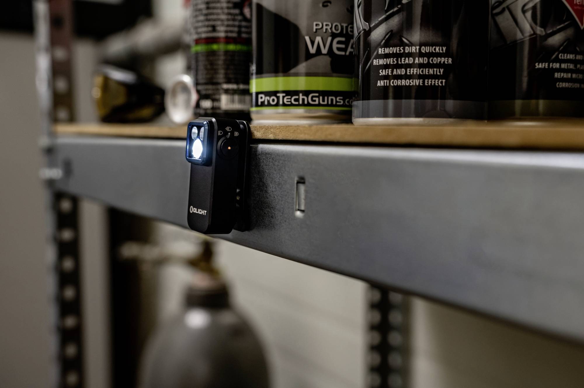 Kleine Taschenlampe mit eingeschaltetem Licht, befestigt an einem Metallregal in einer Werkstatt. Im Hintergrund Pflegeprodukte für Waffen.