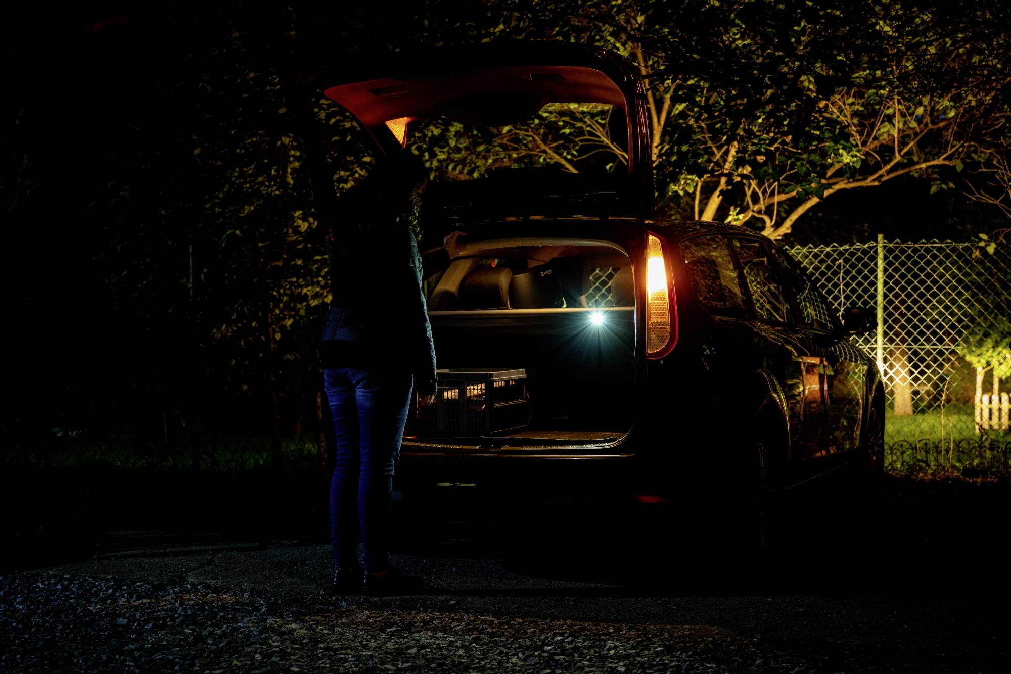 Eine Person steht bei Nacht neben einem Auto mit geöffneter Heckklappe. Der Kofferraum ist beleuchtet. Im Hintergrund ist ein Zaun erkennbar.