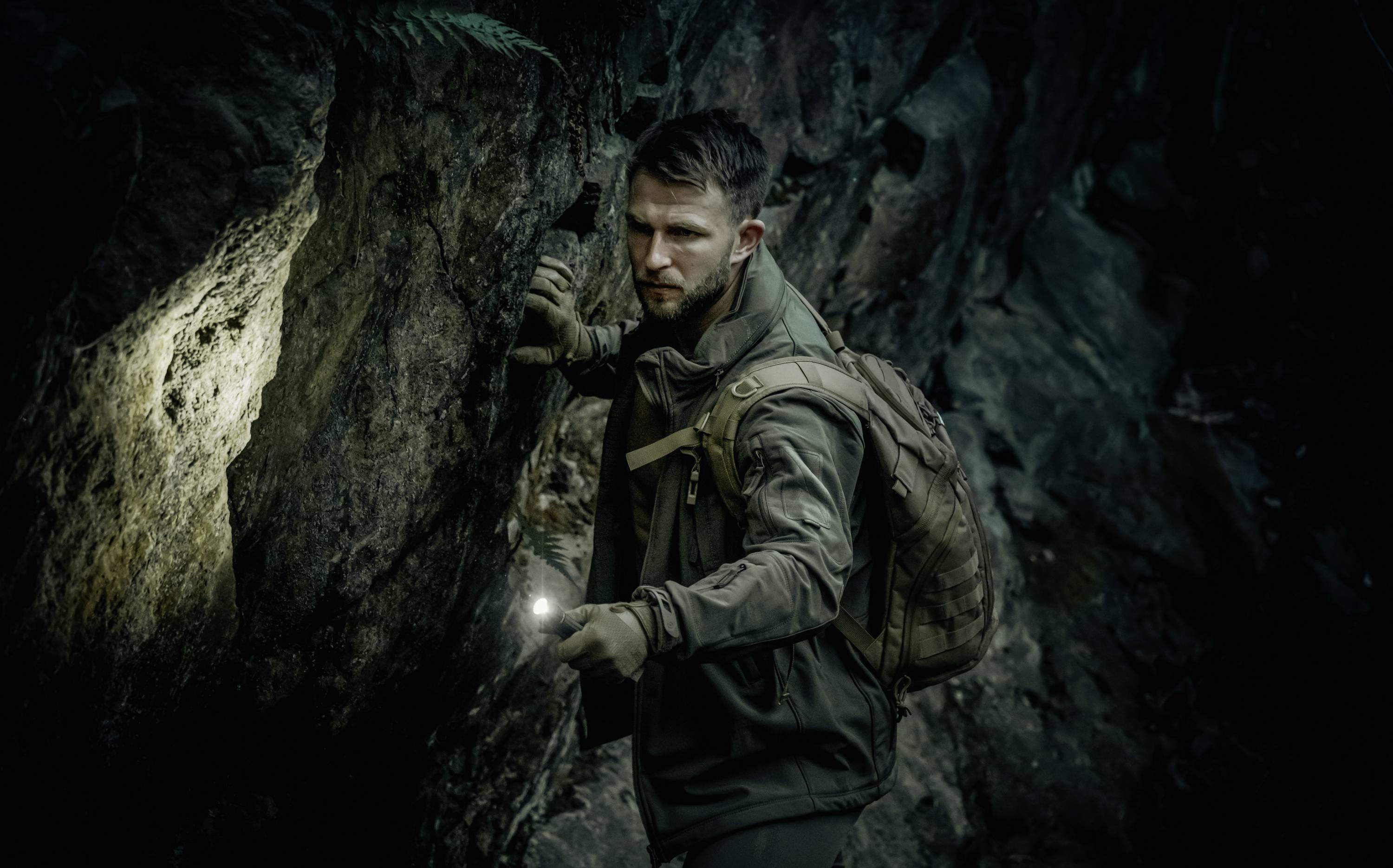 Mann im Wald hält eine Taschenlampe, trägt Outdoor-Kleidung und Rucksack, schaut aufmerksam, umgeben von dunklen Felsen.