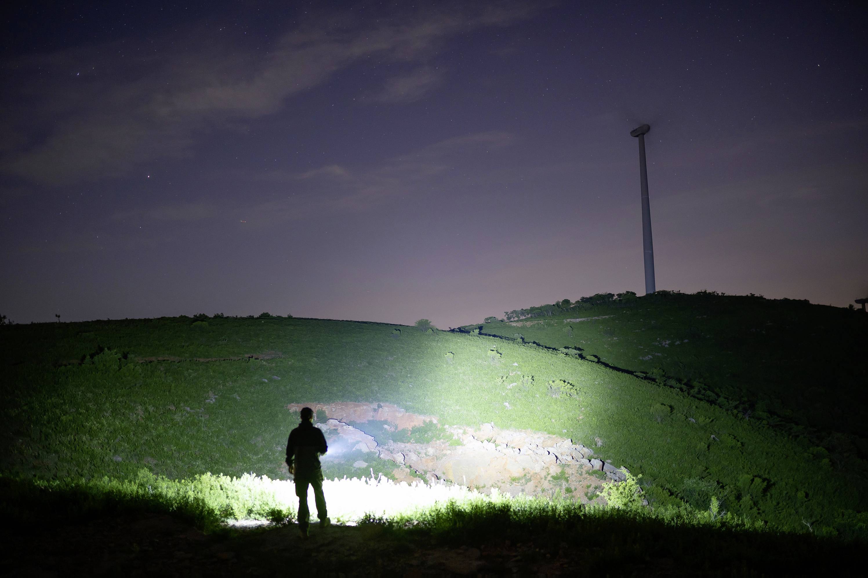 Eine Person steht nachts mit einer Taschenlampe in der Hand auf einem Hügel. Im Hintergrund ist ein Windrad zu sehen.