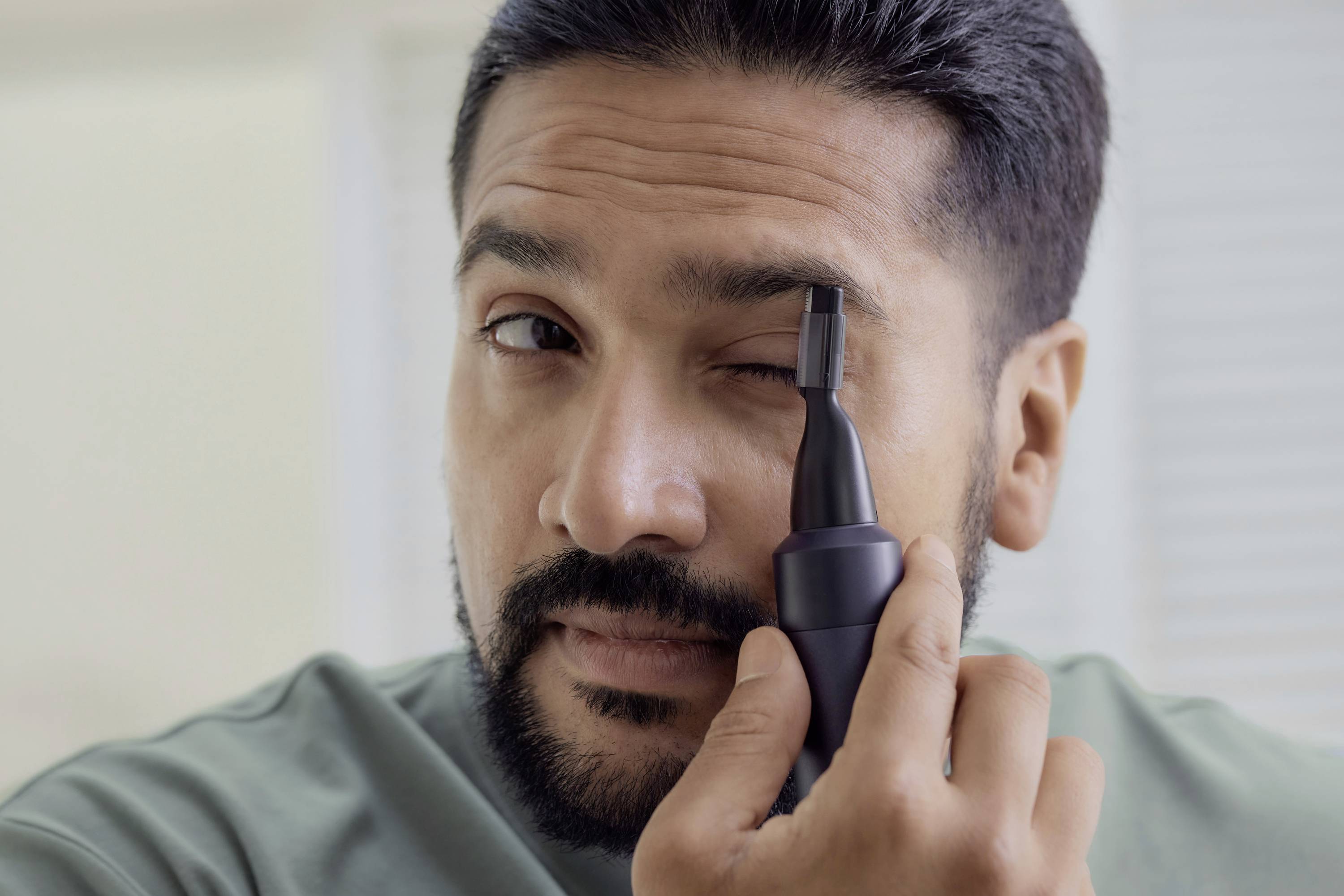 Ein Mann pflegt seine Augenbrauen mit einem elektrischen Trimmer. Er konzentriert sich darauf, präzise zu schneiden.