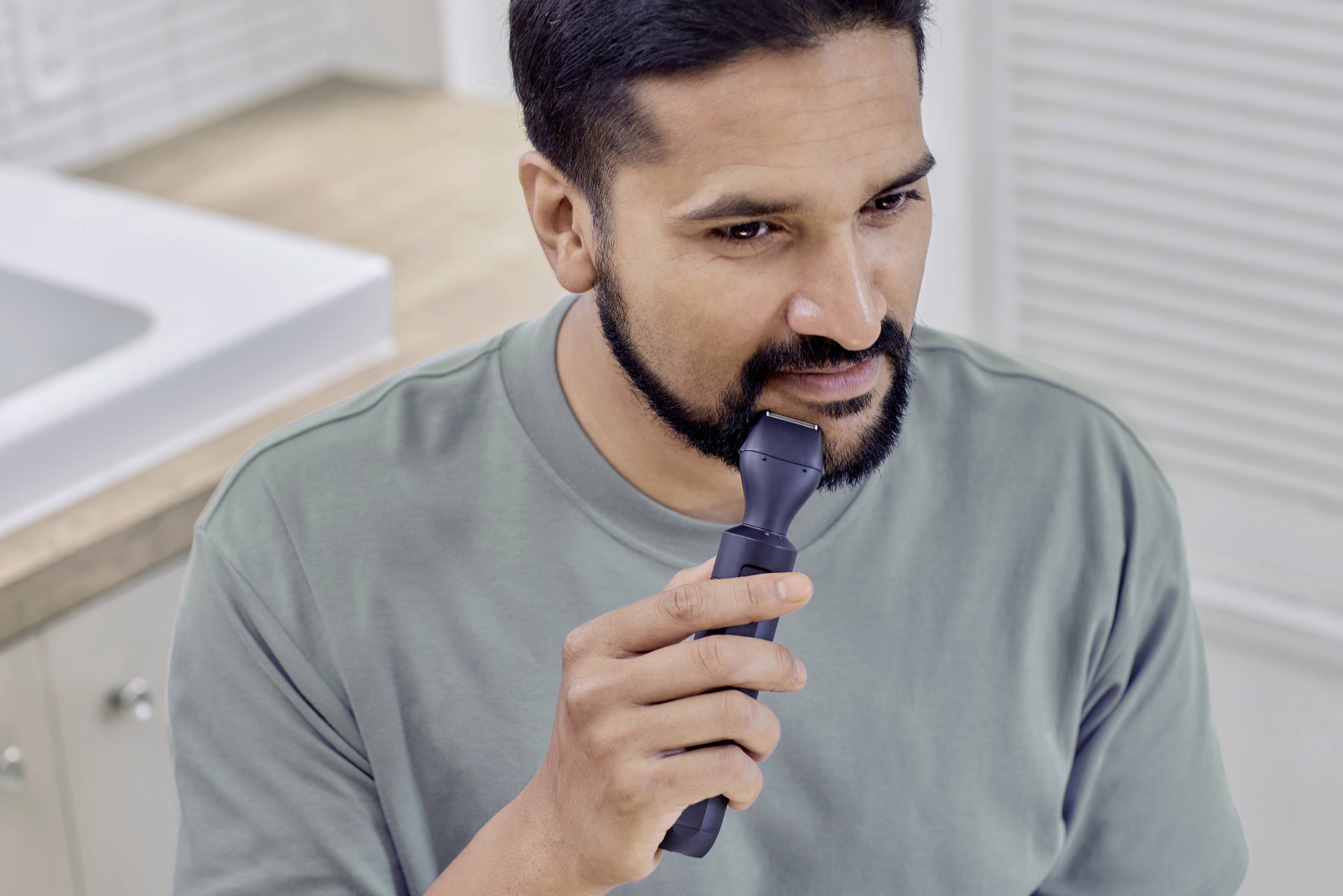 Ein Mann trimmt seinen Bart mit einem elektrischen Trimmer im Badezimmer. Er trägt ein grünes T-Shirt und konzentriert sich auf den Spiegel.