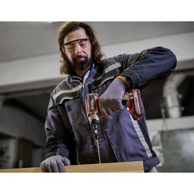 Ein Mann in Schutzbrille und Handschuhen bohrt mit einer elektrischen Bohrmaschine ein Loch in ein Holzstück in einer Werkstatt.