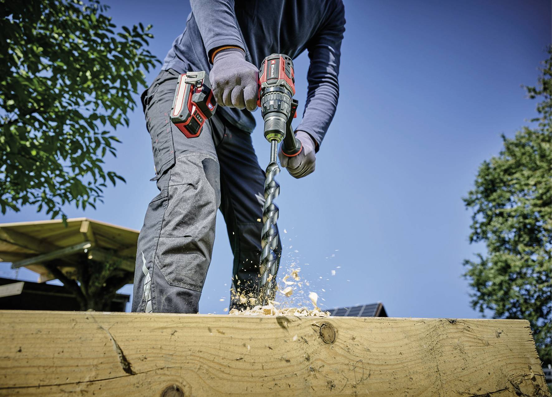Eine Person bohrt mit einer elektrischen Bohrmaschine in Holz. Holzspäne fliegen herum. Es ist ein sonniger Tag im Freien.