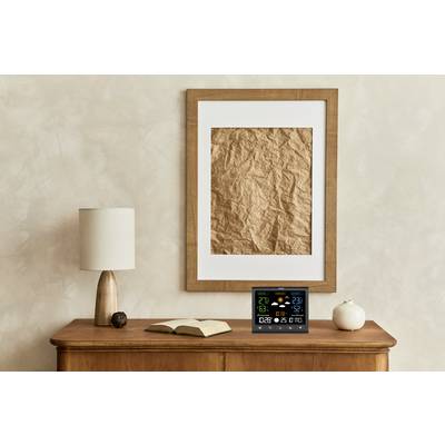 Ein Holzschrank mit einer Lampe, einem aufgeschlagenen Buch, einer kleinen Kürbisfigur und einem digitalen Wetter-Display. Im Hintergrund hängt ein Kunstwerk an der Wand.