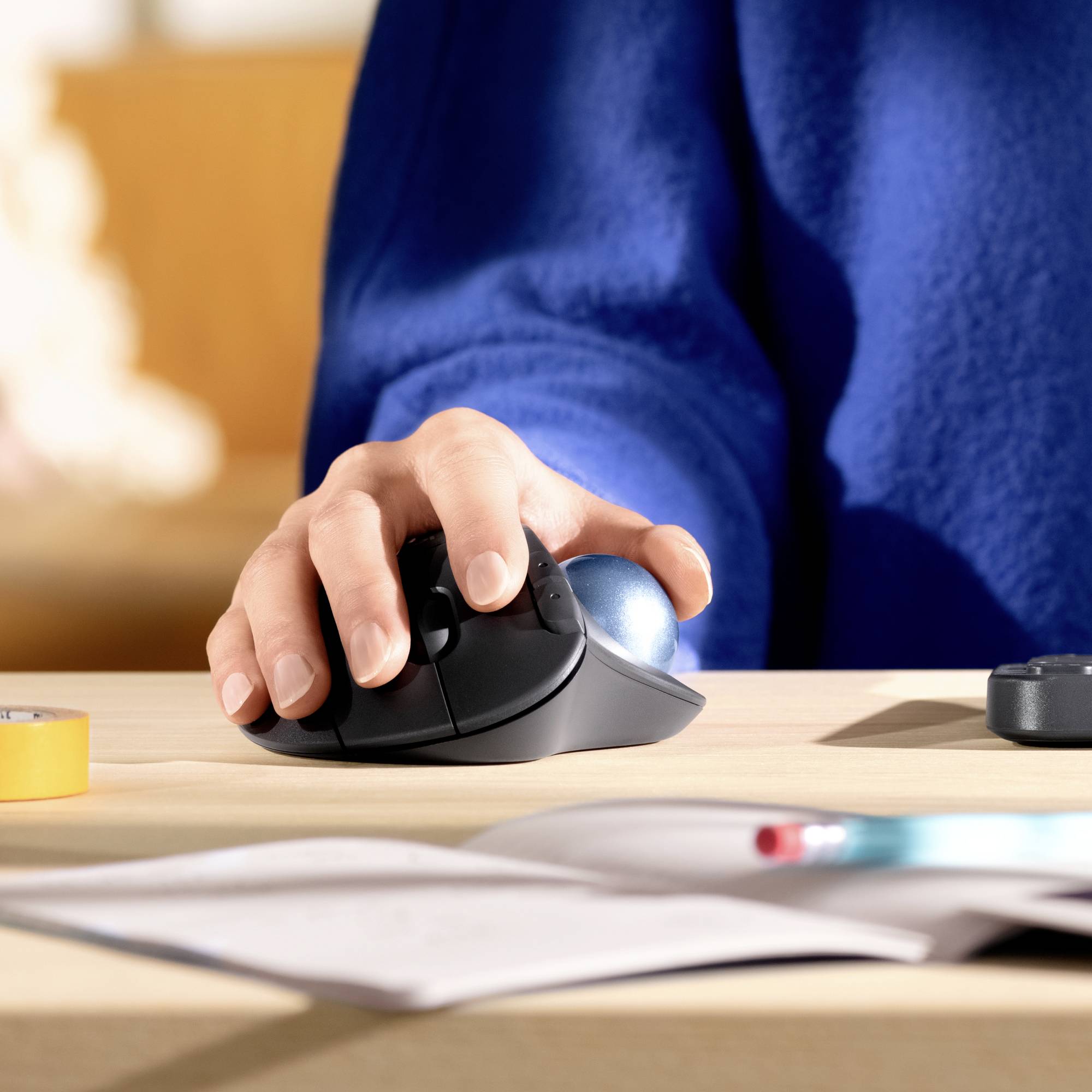 Hand bedient ergonomische Computermaus auf Schreibtisch, umgeben von Papier, Stift und kleinem gelben Klebeband.