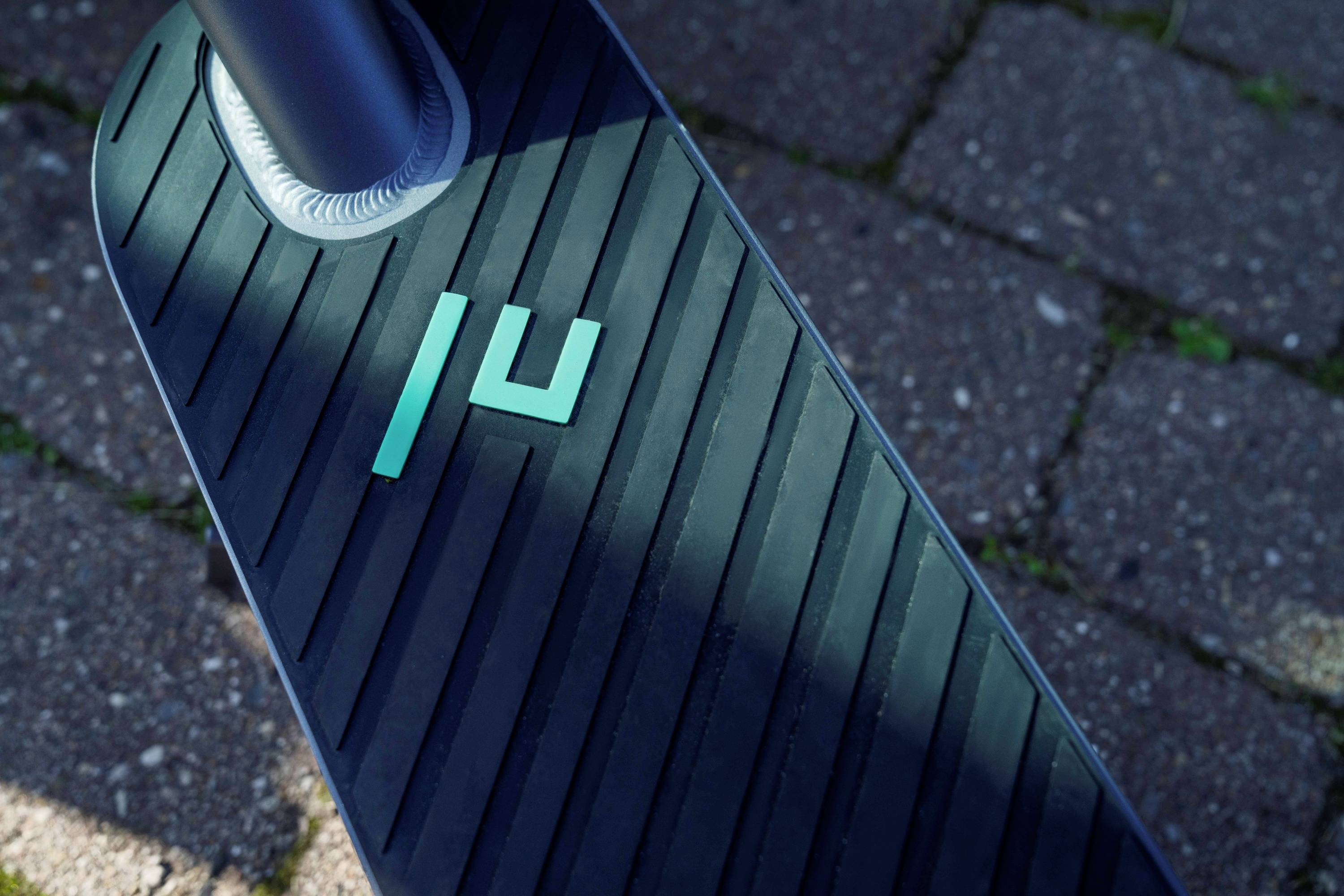 Nahaufnahme eines schwarzen Trittbretts eines E-Scooters mit blauen Streifen und Logo. Darunter Pflastersteine eines Gehwegs.