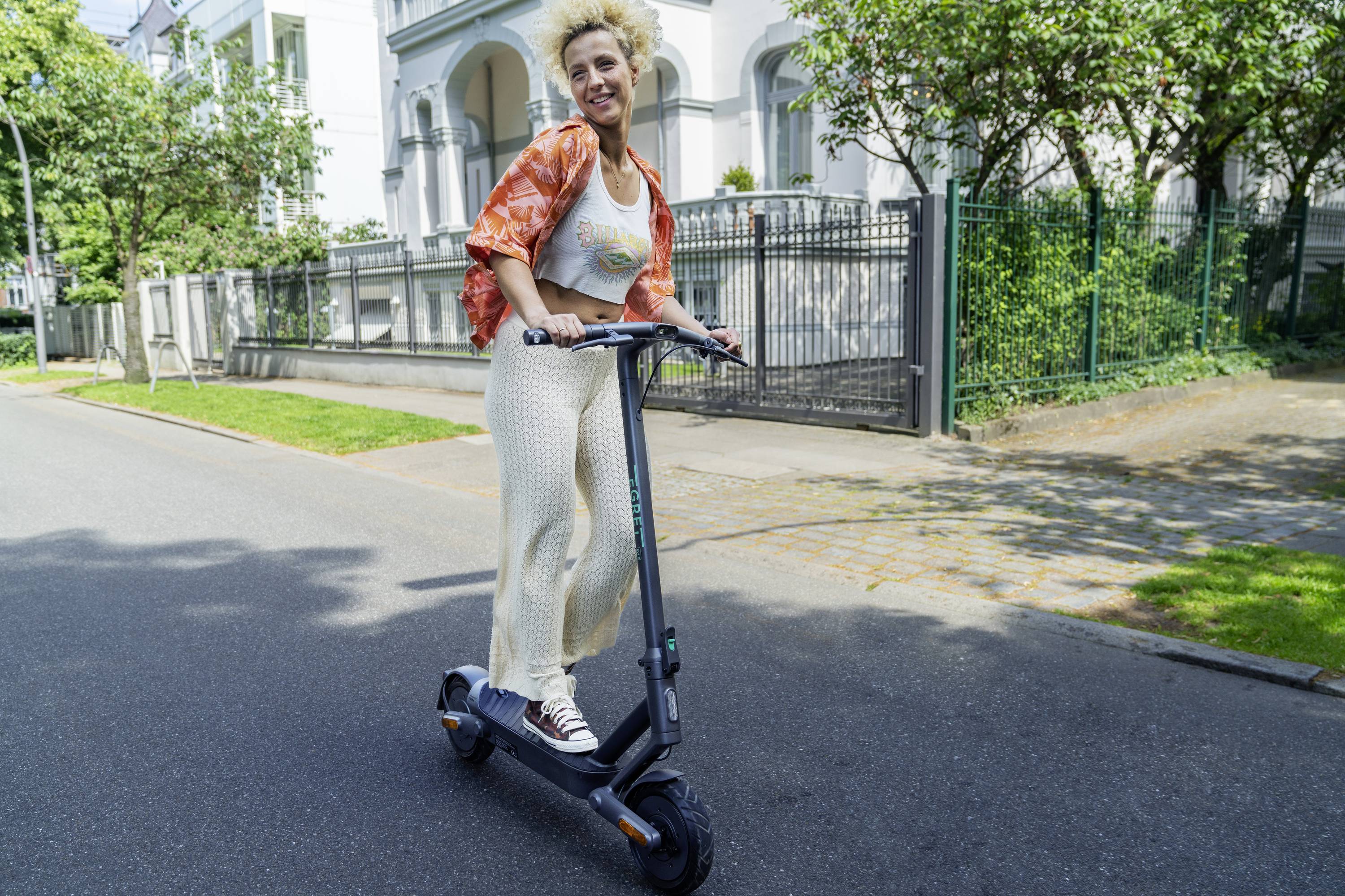 Frau fährt auf einem E-Scooter auf einer sonnigen Straße. Sie trägt eine orangefarbene Jacke, lächelt und wirkt entspannt.