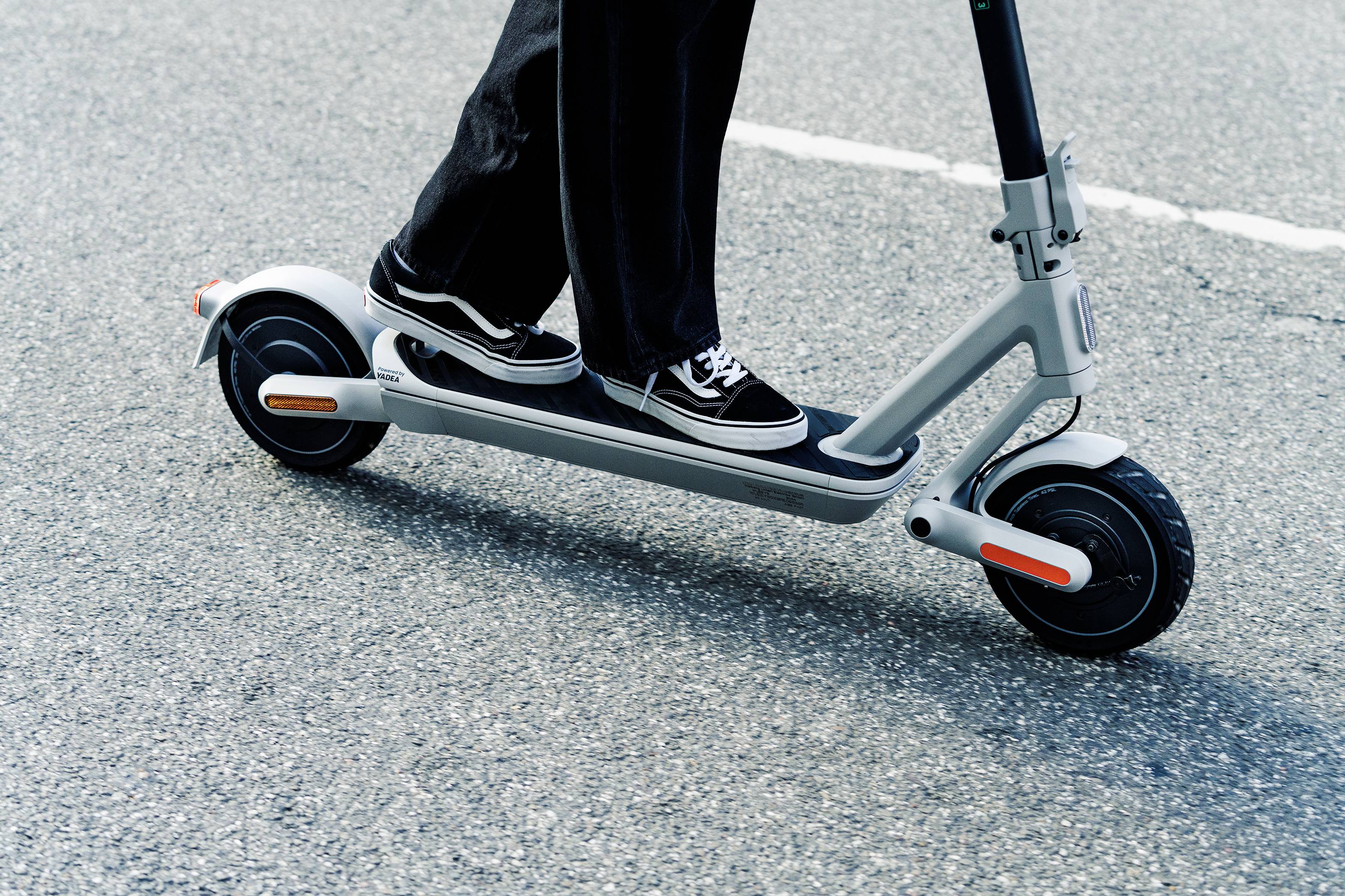 Person fährt auf einem elektrischen Tretroller über den Asphalt. Der Roller ist grau mit schwarzen Reifen und orangen Akzenten.