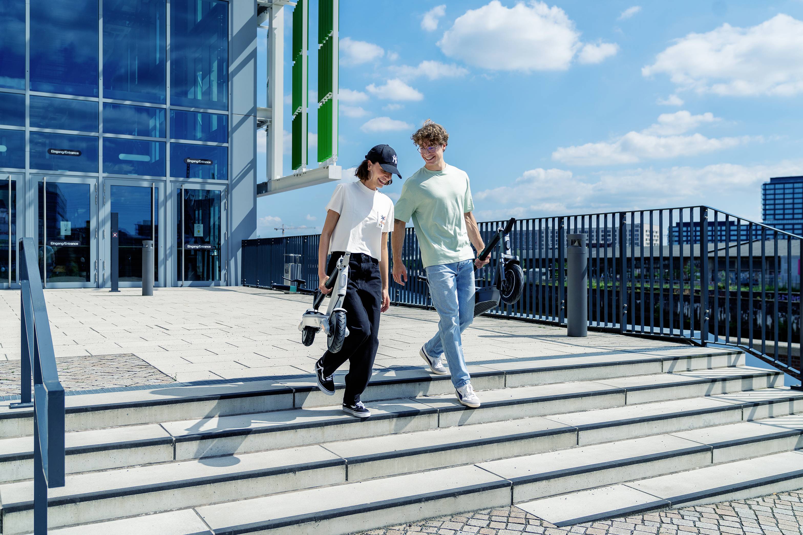 Zwei Personen gehen eine Treppe vor einem modernen Gebäude hinunter, tragen lässige Kleidung und unterhalten sich bei sonnigem Wetter.