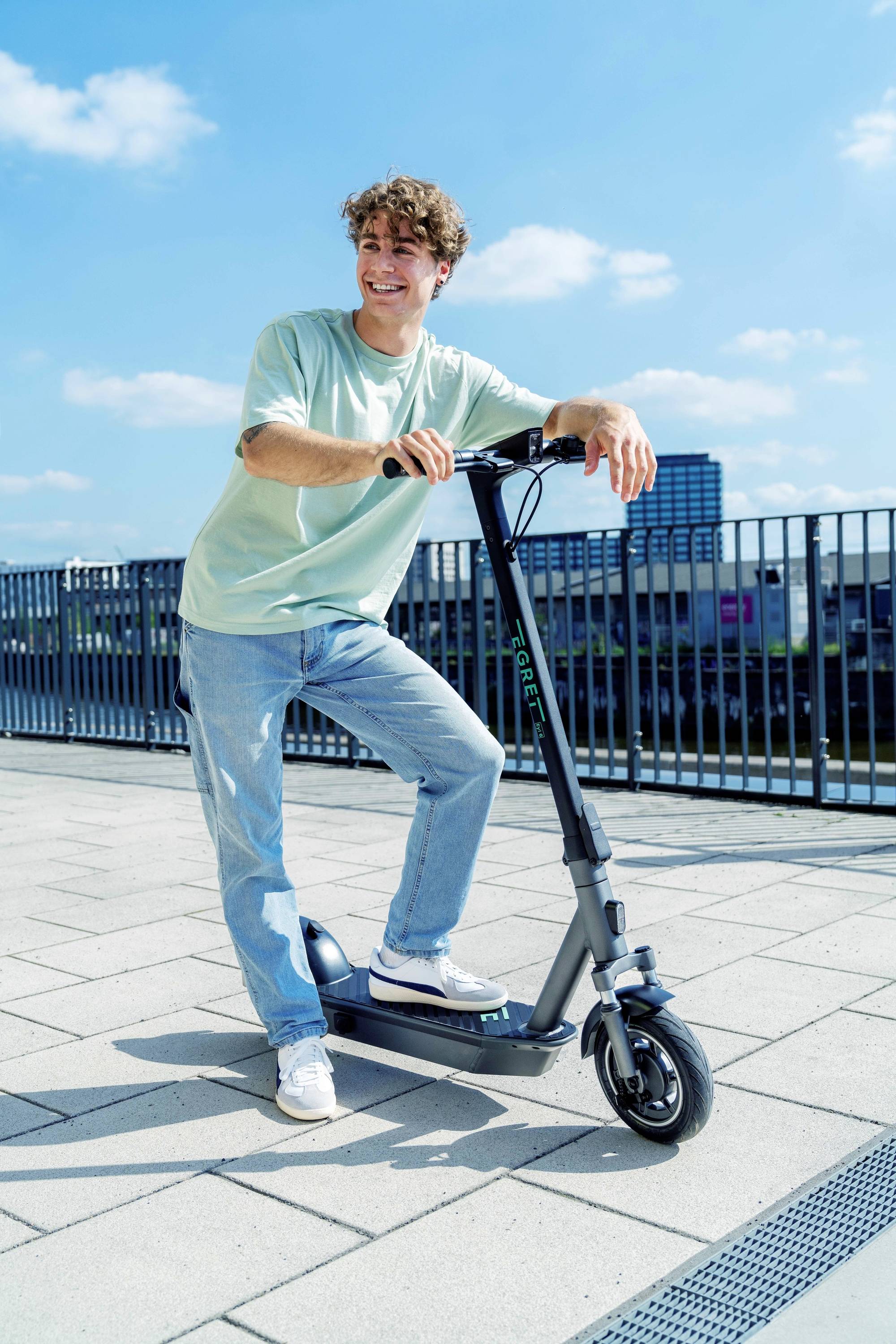 Ein junger Mann steht lächelnd auf einem Gehweg und lehnt sich an einen E-Scooter. Im Hintergrund sind ein Zaun und Gebäude zu sehen.