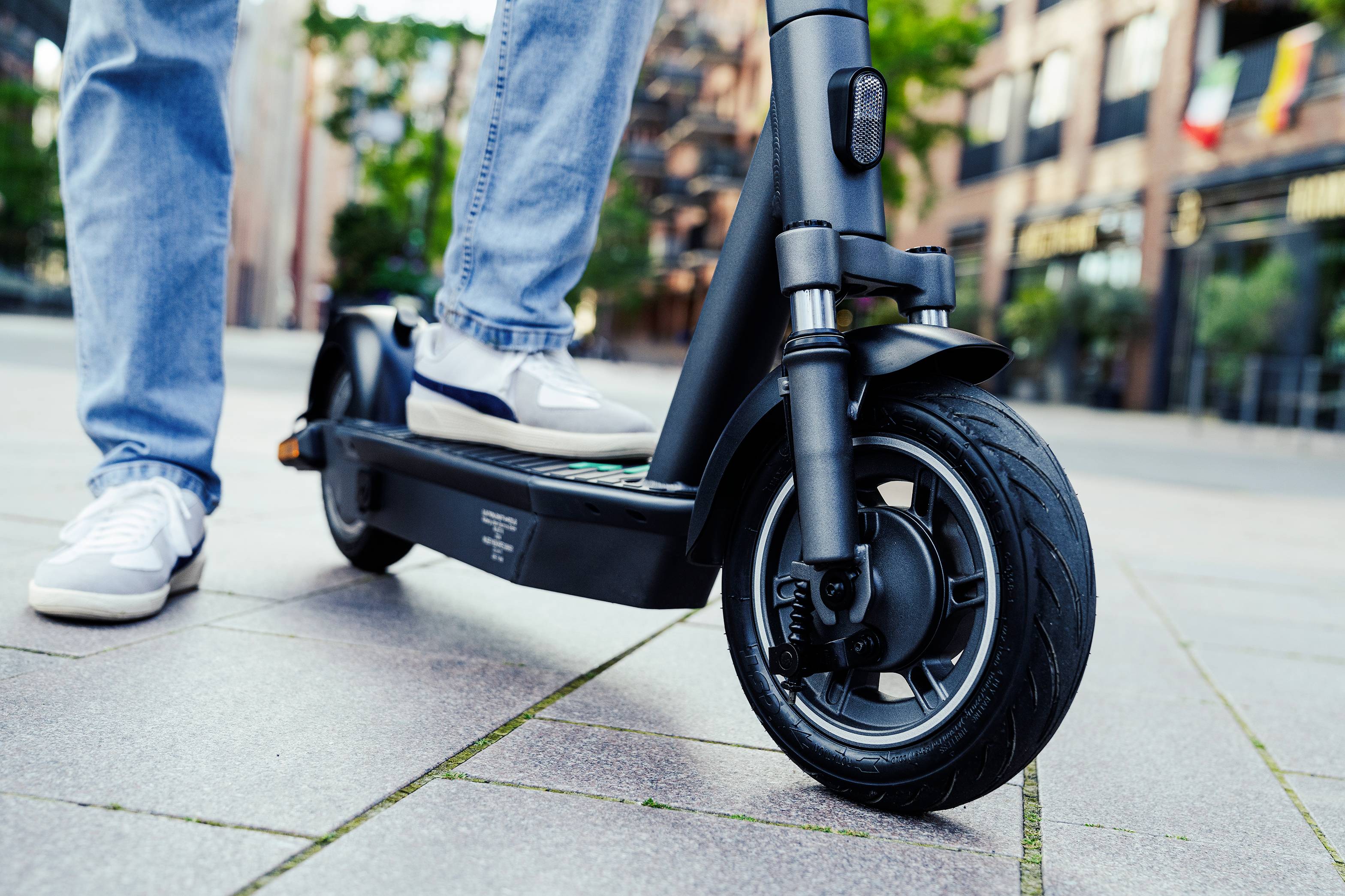 Eine Person steht auf einem elektrischen Scooter auf einer gepflasterten Stadtstraße. Im Hintergrund sind Gebäude und Fahnen zu sehen.