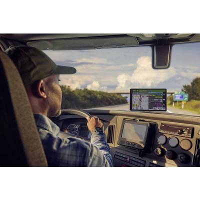 Ein Mann fährt einen Lkw auf einer Landstraße, schaut auf ein Navigationsgerät auf dem Armaturenbrett. Die Landschaft ist sonnig.