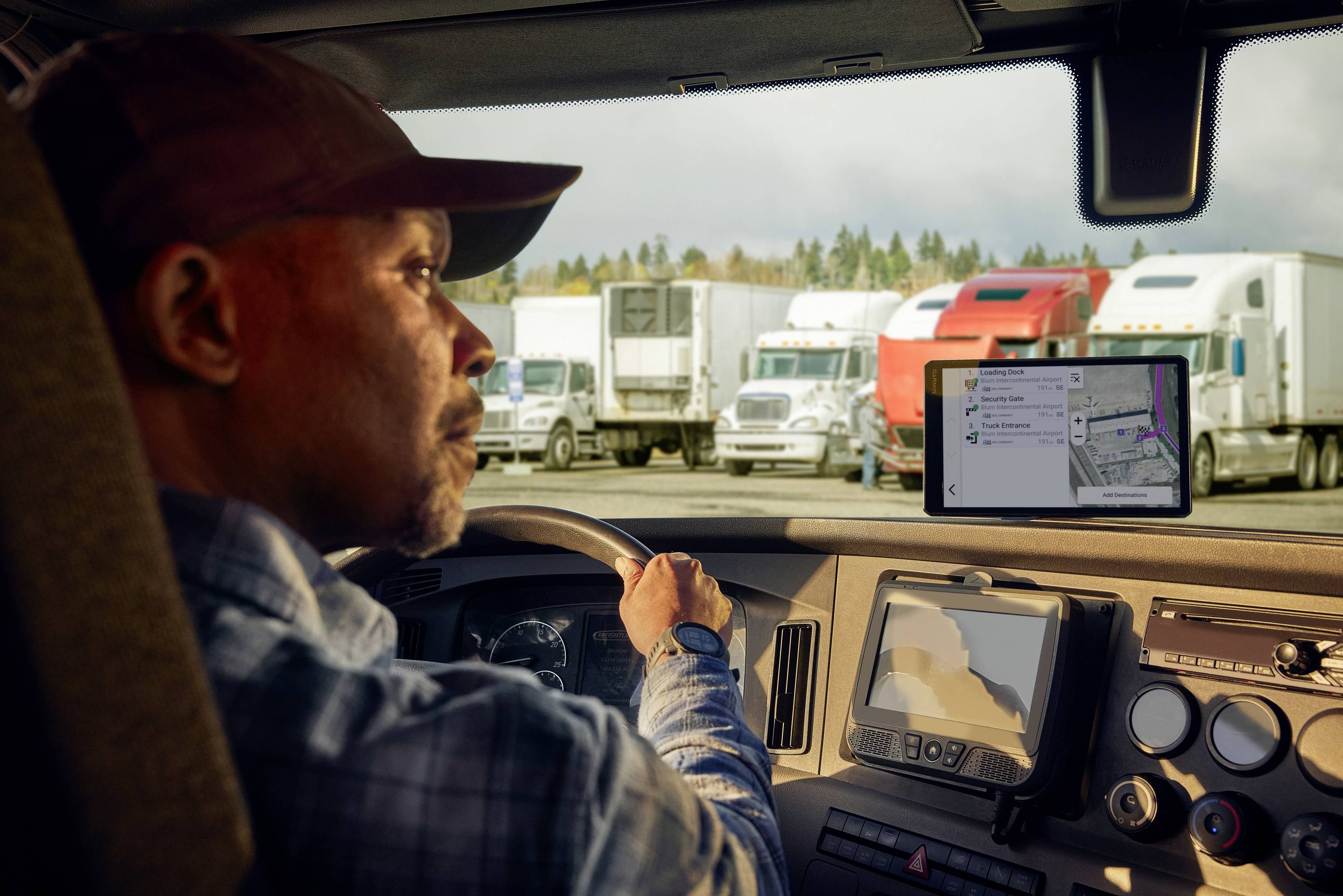 Ein Lkw-Fahrer sitzt in einem Lkw und schaut auf ein angeschaltetes Navigationsgerät, während mehrere geparkte Lkws im Hintergrund zu sehen sind.
