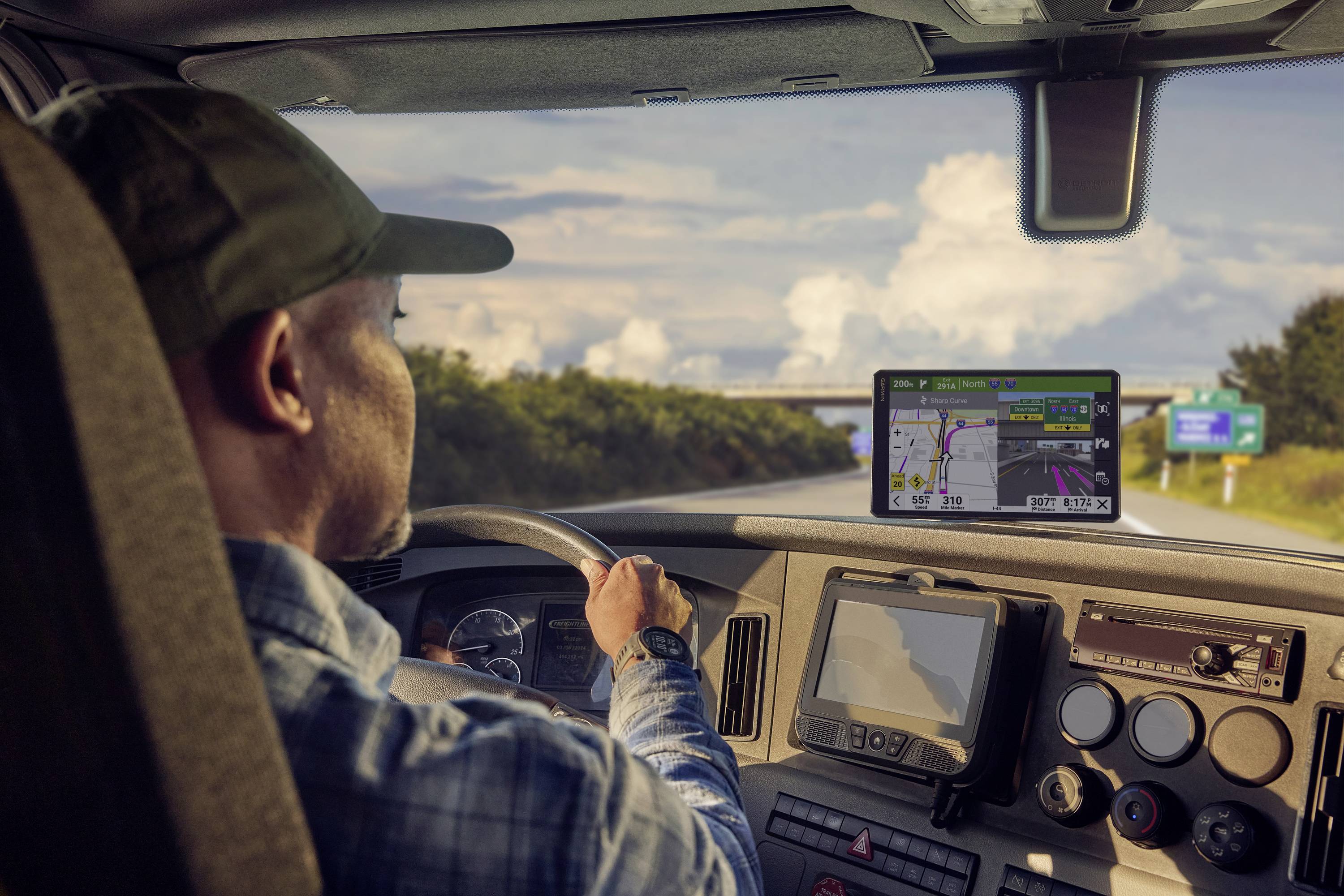 Ein Fahrer in einem LKW blickt auf ein GPS-Navigationsgerät, das eine Route auf der Autobahn anzeigt. Himmel mit Wolken im Hintergrund.