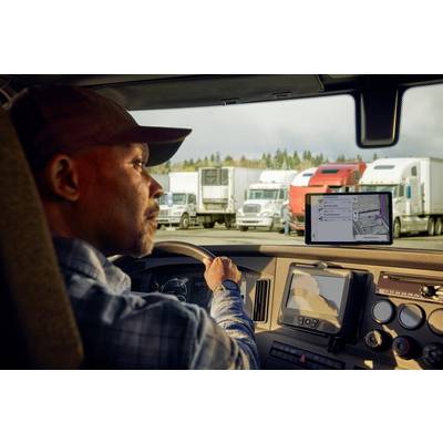 Ein Lkw-Fahrer sitzt in der Fahrerkabine und betrachtet ein Navigationsgerät. Im Hintergrund stehen mehrere geparkte Lastwagen.