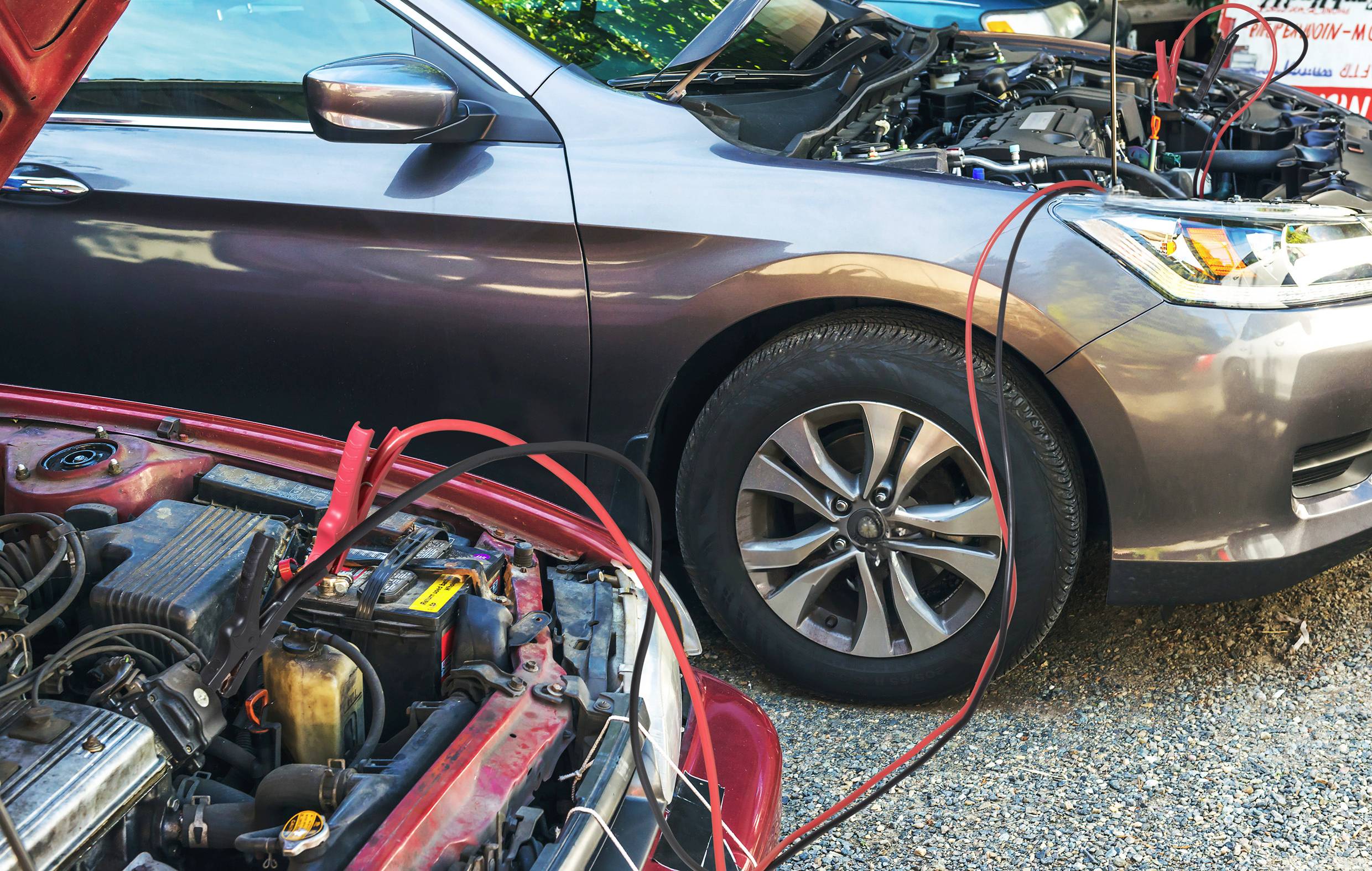 Zwei Autos stehen mit geöffneter Motorhaube, verbunden durch Überbrückungskabel, was auf das Überbrücken einer Autobatterie hindeutet.