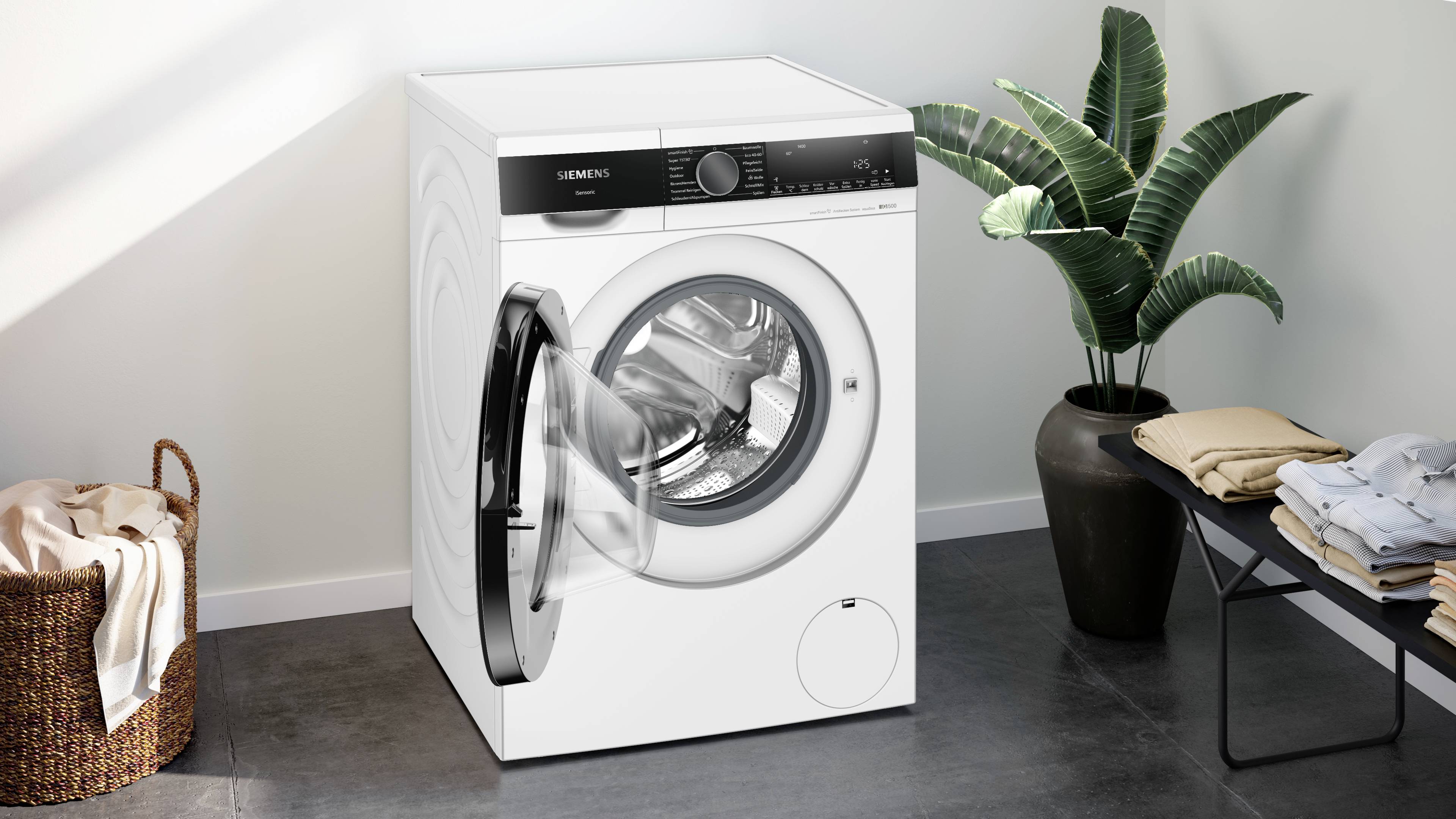 Eine moderne, weiße Waschmaschine in einem hellen Raum, umgeben von einem Korb mit Wäsche und einer Pflanze.